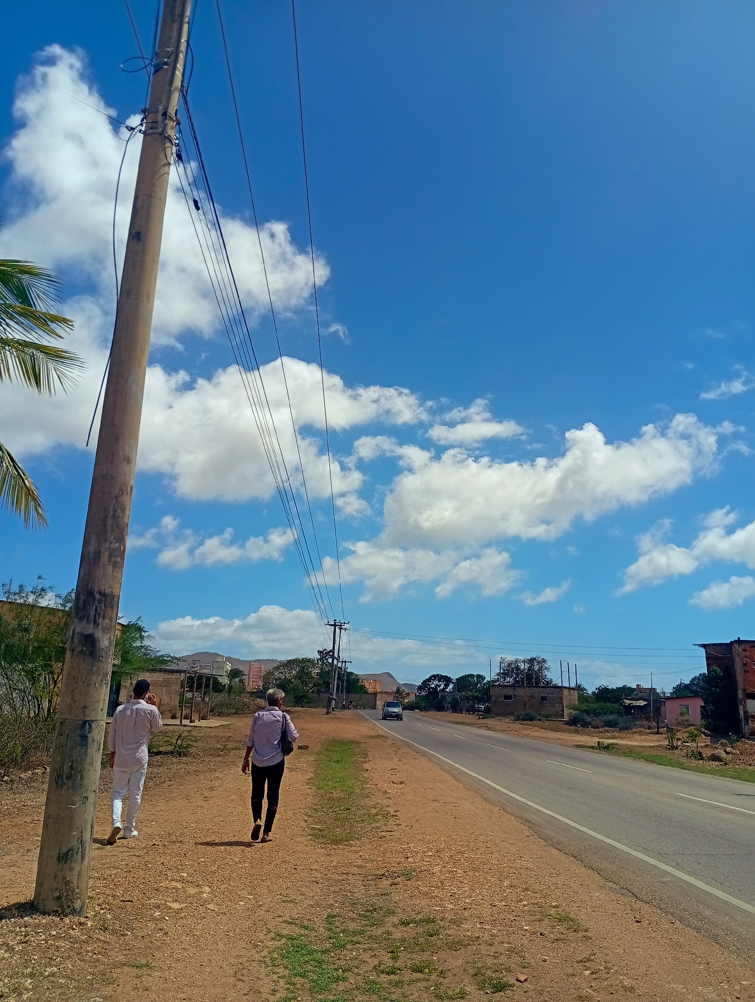 The image depicts a bright, sunny day in a rural or semi-rural area, likely on the outskirts of Juan Griego, Venezuela. The foreground features a dirt and gravel path alongside a paved road. A tall, weathered utility pole with numerous wires runs vertically along the left side of the image. Two men are walking away from the camera on the path. The man on the left wears a light-colored collared shirt and white pants, while the man on the right wears a light-colored long-sleeved shirt and dark pants. Both are walking in the same direction, towards the road.
The road curves gently to the right and is bordered by a sparse, dry grassy verge. On the right side of the road, there are various buildings, including what appear to be low-rise, possibly unfinished structures and a small pink house. Further in the distance, a car is visible driving on the road. The background consists of rolling hills and more vegetation. The sky is a vibrant blue with scattered, fluffy white clouds. The overall impression is one of a typical day in a warm climate, possibly a developing area.