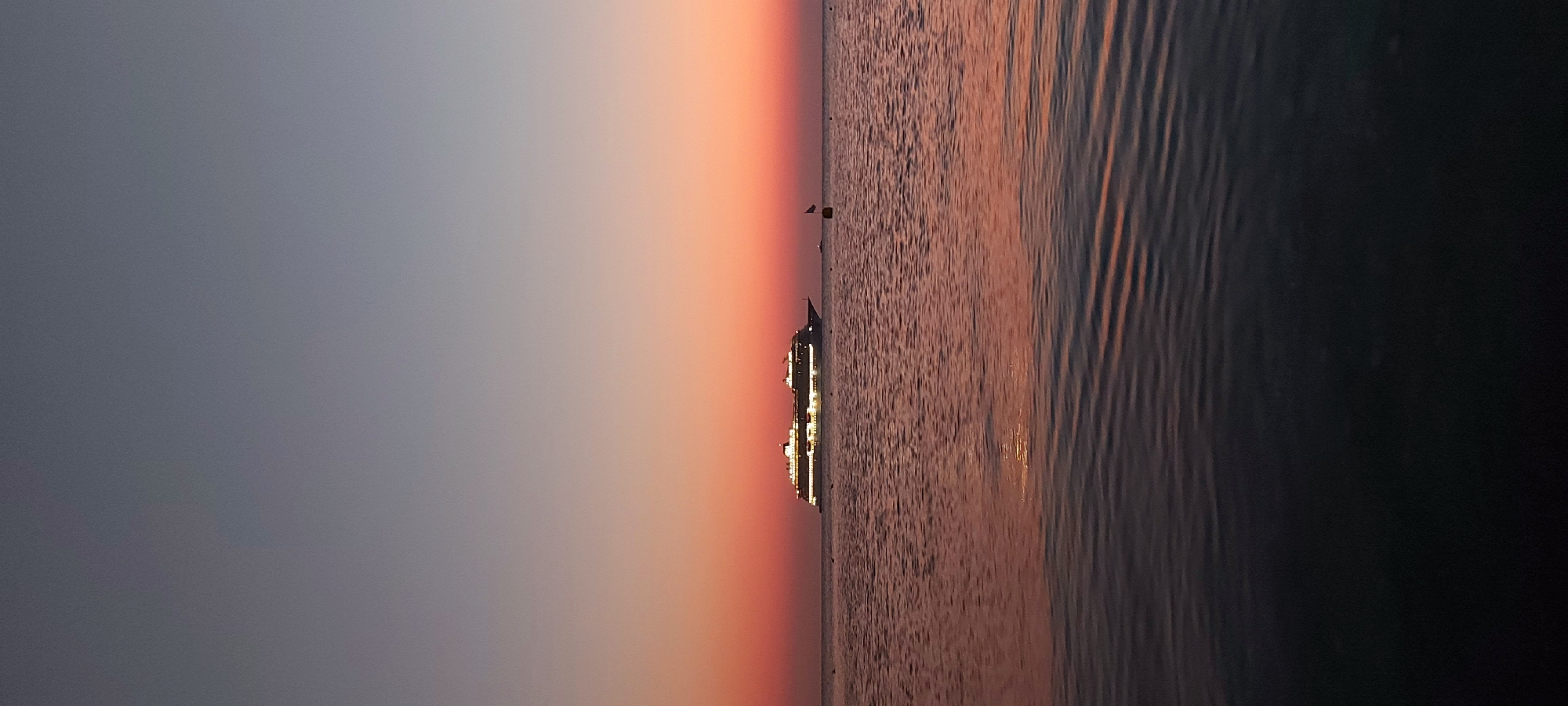 A long cruise ship is illuminated and anchored on the calm sea during twilight. The sky is a gradient of pale blue at the top transitioning to soft orange and pink hues near the horizon. The water reflects the colors of the sky and the ship's lights. A small buoy with a red flag is visible in the distance to the right of the ship. The scene is devoid of people and shows no signs of activity beyond the anchored vessel. The location is the Municipal Unit of Rhodes, Greece.