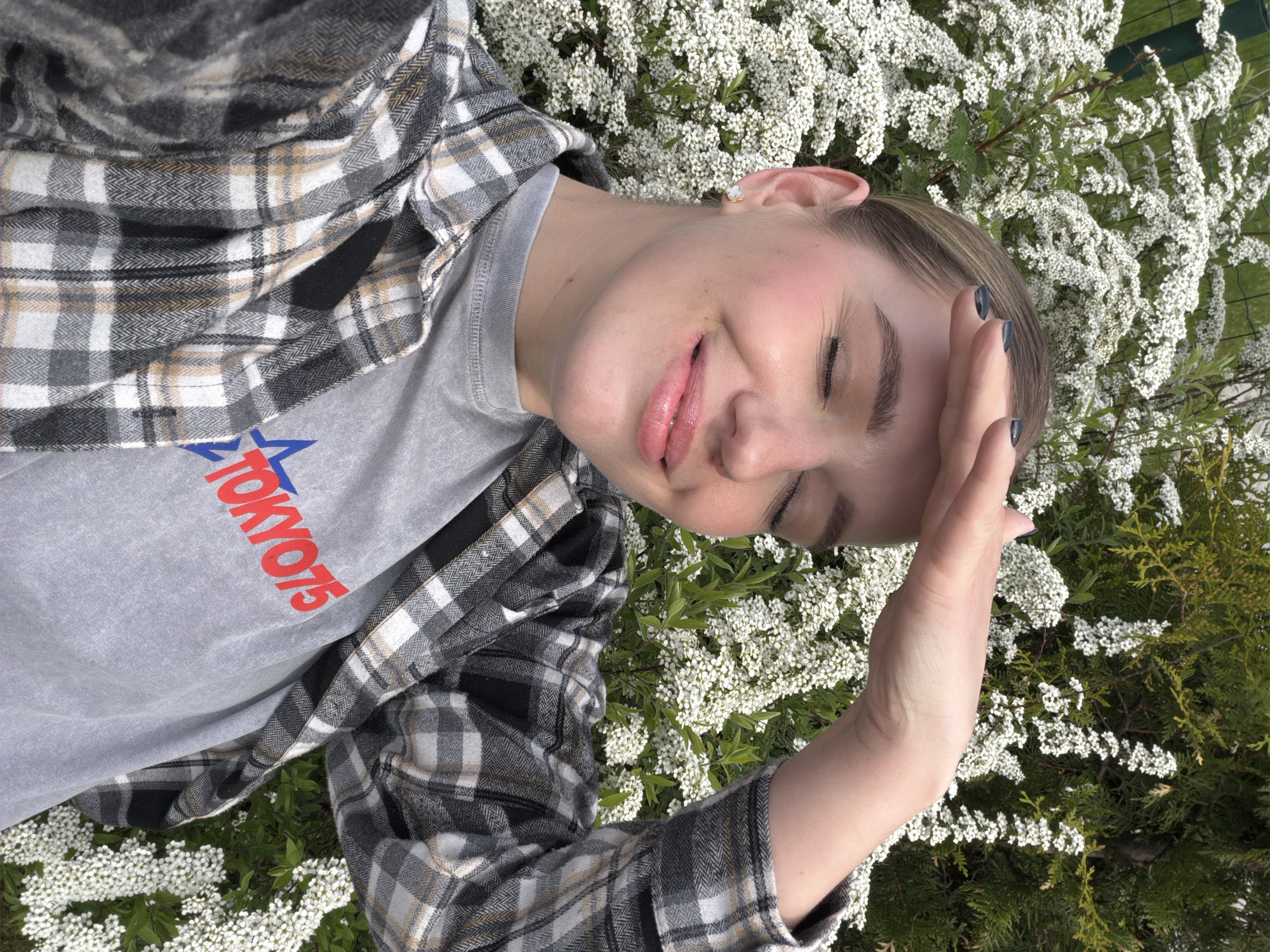 A young woman is lying on a bed of small white flowers. She is wearing a gray T-shirt with the word "TOKYO" written in red and a blue star. Over the T-shirt, she has on a gray and white plaid flannel shirt. Her hand is raised to her forehead, possibly shielding her eyes from the sun. Her eyes are closed. The photo appears to be taken outdoors with a mix of white flowers and some greenery in the background. The location cannot be determined from the image provided.