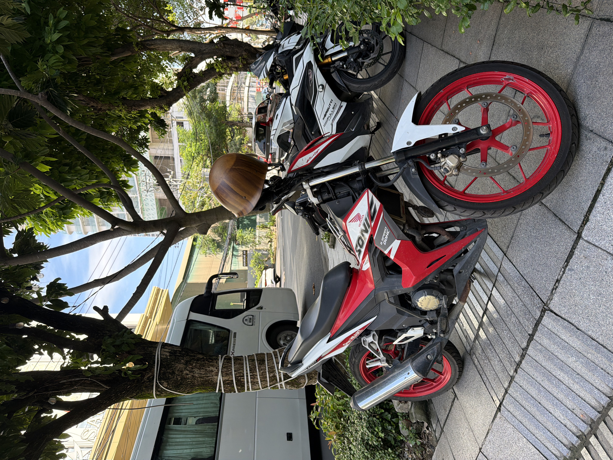 A Honda Sonic motorcycle, predominantly red and white with black accents, is parked on a sidewalk in Nha Trang, Vietnam. The motorcycle is in the foreground, angled slightly towards the viewer's left. A wooden-colored helmet is placed on its handlebars. Behind the Honda Sonic, a white and black motorcycle is parked, partially obscured by green foliage on the right. In the background, a white bus is parked to the left, with its rear section visible. Further back, a multi-story building and more trees line the street. The sidewalk is paved with grey tiles, some with textured, ribbed surfaces. The overall scene depicts a typical urban streetscape in Vietnam.