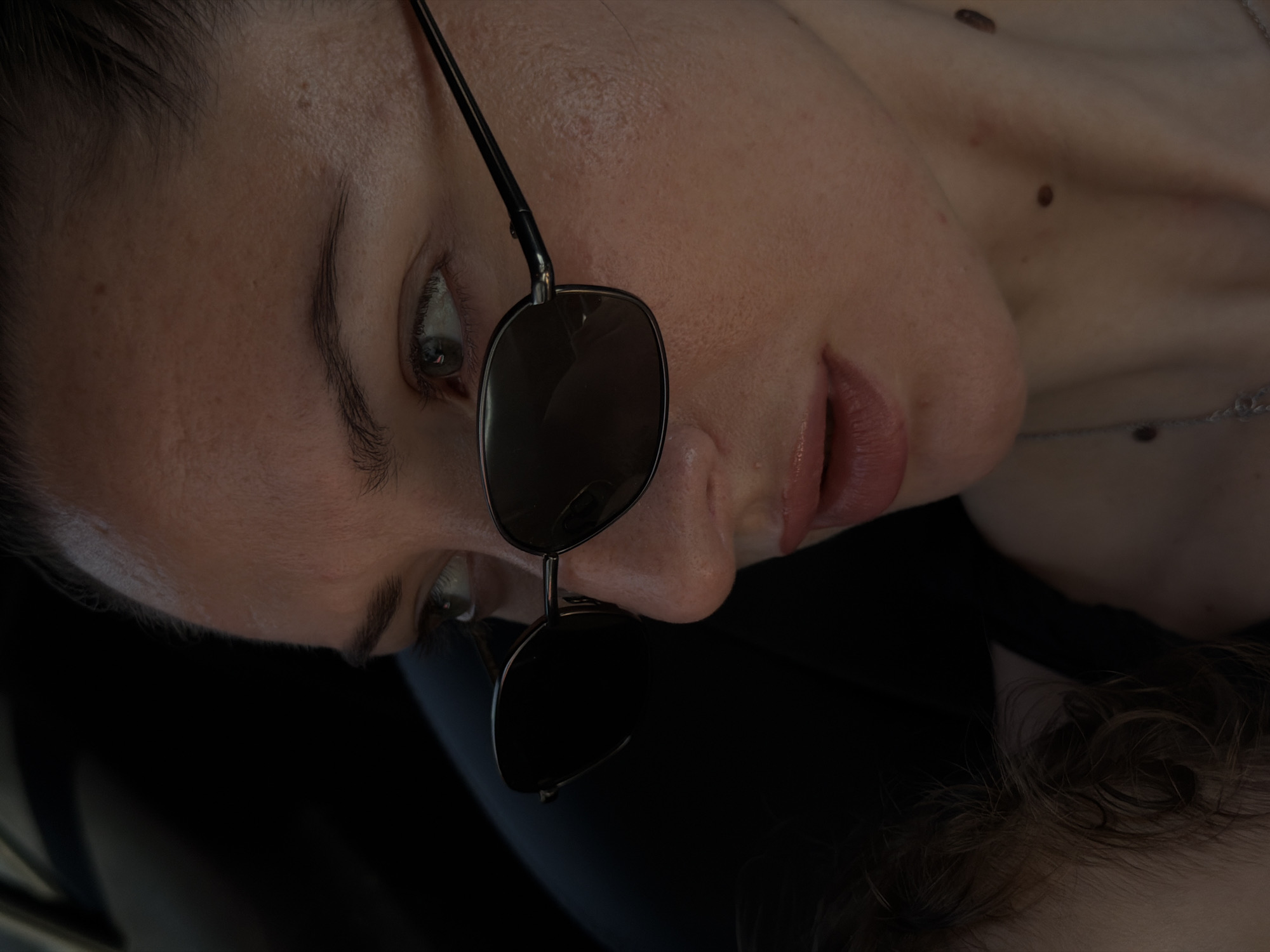 The image is a close-up of a woman wearing sunglasses, her face angled to the side and her mouth slightly open. Her skin has visible texture and some moles. A dark, out-of-focus area on the left suggests a background, possibly the interior of a vehicle. The lighting is dramatic, with strong highlights and shadows. No elements in the image directly indicate a location in Sharm El Sheikh, Egypt.
