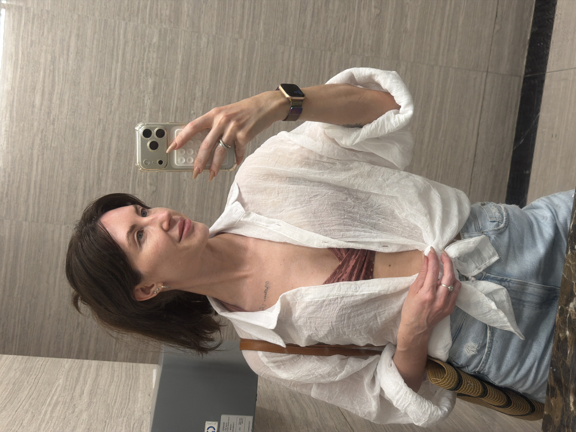 This image depicts an adult female capturing a mirror selfie in an indoor public setting, likely a restroom or changing area, located in Nha Trang Ward, Vietnam.
The woman has medium-dark brown hair, casually styled, and a fair complexion. She is smiling slightly and looking to her right (viewer's left). Her left ear features three small earrings: two studs and one small hoop. She has a faint script tattoo on her upper chest, below the collarbone, and another visible on her right forearm.
She is wearing a white, sheer, textured long-sleeve button-up shirt, unbuttoned and tied at the waist, revealing a burnt orange/terracotta-colored textured bralette or bikini top underneath. Her lower garment consists of light blue denim shorts with distressed detailing.
On her right hand, she holds a silver-toned smartphone with a clear case and a white rectangular grip accessory, positioning it for the selfie. Her right ring finger bears a double-banded ring, and her fingernails are long, almond-shaped, and painted with a neutral polish. An Apple Watch-style smartwatch with an iridescent metallic mesh band is visible on her right wrist. Her left hand is positioned near the knot of her shirt, and she wears a single-stone ring on her left ring finger. A woven straw-like bag with brown and black stripes and a brown strap is slung over her left shoulder.
The background consists of light brown/grey, horizontally striated large format tiles on the walls. To the left, a dark grey wall-mounted dispenser with a white label is visible. The foreground shows a dark, reflective surface, likely a countertop or mirror edge, and the floor features similar light brown/grey tiles with a distinct dark horizontal tile strip at the base.
