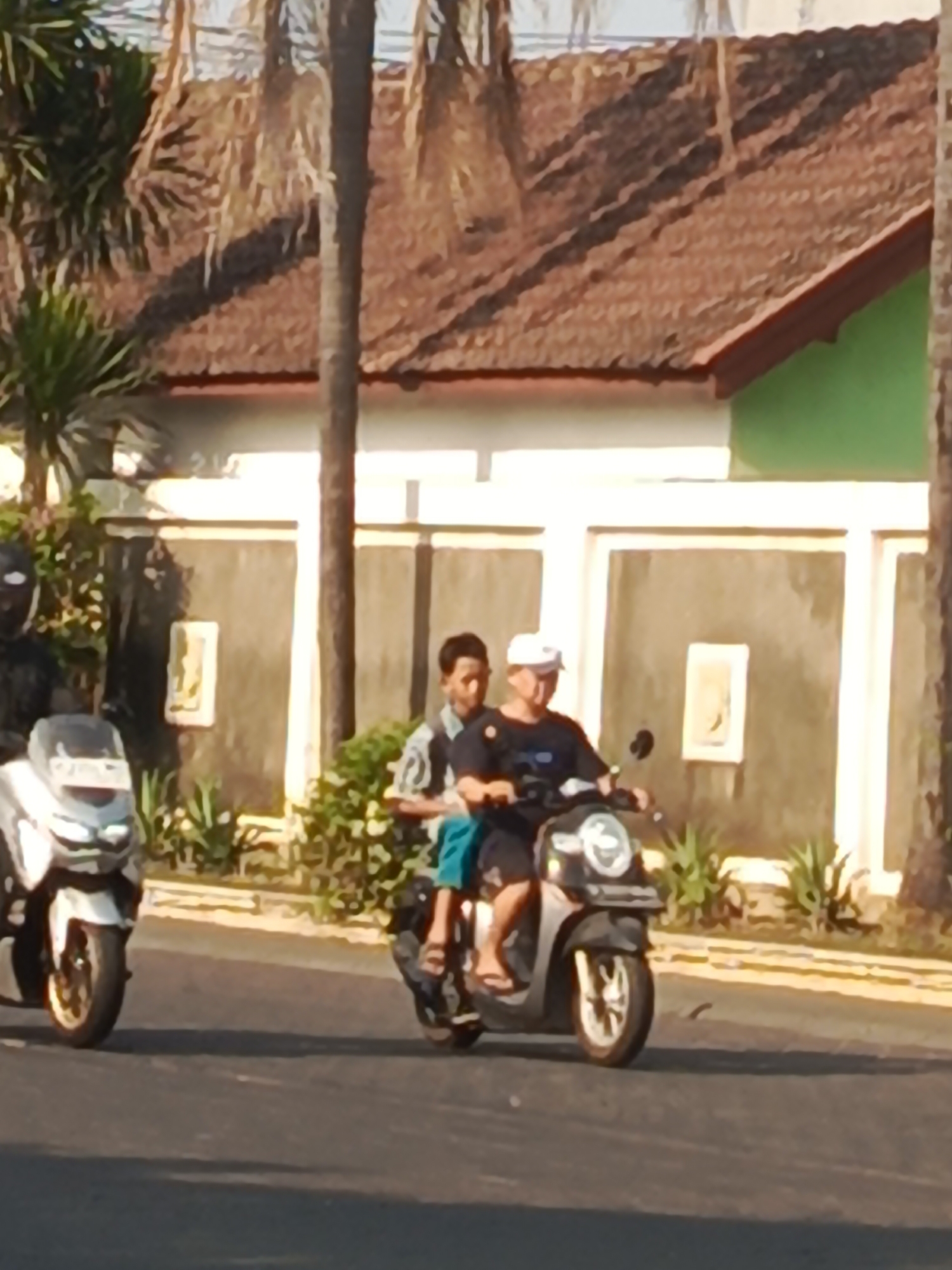 In Boro, Indonesia, two individuals are riding on a motorcycle down a street. The rider in front wears a white cap and a dark t-shirt, while the passenger behind him has short hair and is wearing a light-colored shirt and blue shorts. The motorcycle is dark-colored with silver accents. To the left of the motorcycle, another person is visible on a white scooter, wearing a black helmet and jacket. The background features a building with a tiled roof and a textured wall, lined with plants. The lighting suggests it is daytime.