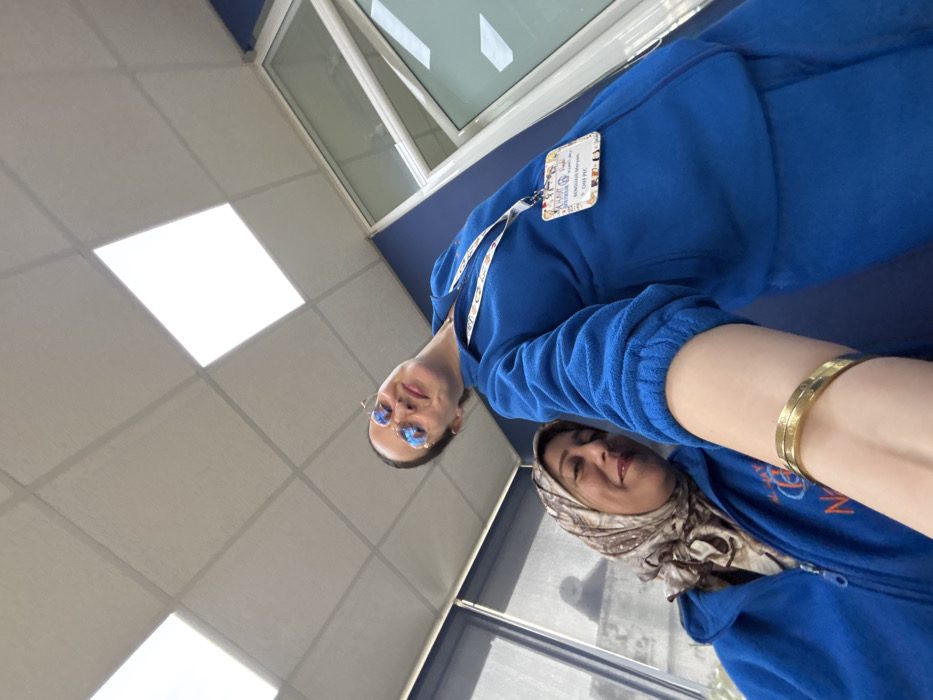 The image depicts two adult females indoors, captured from a low-angle selfie perspective. Both individuals are wearing royal blue fleece zip-up jackets.
The foreground subject, identified by an ID badge on a lanyard as "BENGUAM Maryam, CHEF P.I.C." for "AL HAYAT 6 PREMIUM," is partially obscured by her extended right arm, which wears a gold-colored bangle. She has dark hair, wears round, light-tinted sunglasses, and offers a slight smile.
The second subject, positioned behind and to the left, wears a multi-toned patterned headscarf in shades of brown and beige. Her blue fleece jacket displays a partially visible "AL HAYAT 6 PREMIUM" logo with an additional large "N" below it on the left chest. She is smiling and looking towards the camera.
The environment is an indoor commercial or office space. The ceiling is a white suspended grid system with four large, illuminated rectangular panel lights. The background features a dark blue wall section on the right, adjacent to a white-framed glass partition or window. On the left, a window with vertical blinds allows ambient light into the space.
Based on the identification visible on the ID badge ("AL HAYAT 6 PREMIUM" and "BENGUAM Maryam"), the location is an "Al Hayat 6 Services" facility in Meknes, Morocco, consistent with a professional service center or office.