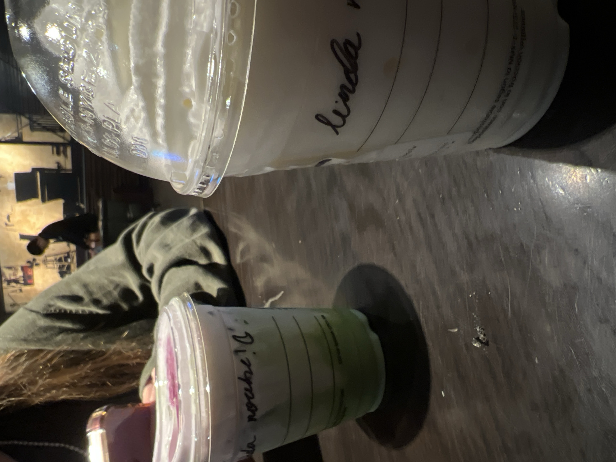 The image shows two Starbucks drinks sitting on a dark, textured surface, likely a table. The larger drink, labeled "linda v", is mostly white with a clear lid containing whipped cream. The smaller drink, labeled "Da noche D", is green at the bottom and white at the top, also with a clear lid.
A person with long brown hair and wearing a green jacket is partially visible in the left side of the frame. The background shows indistinct wall decor, which suggests an indoor location, possibly a Starbucks in Mexico City, Mexico.