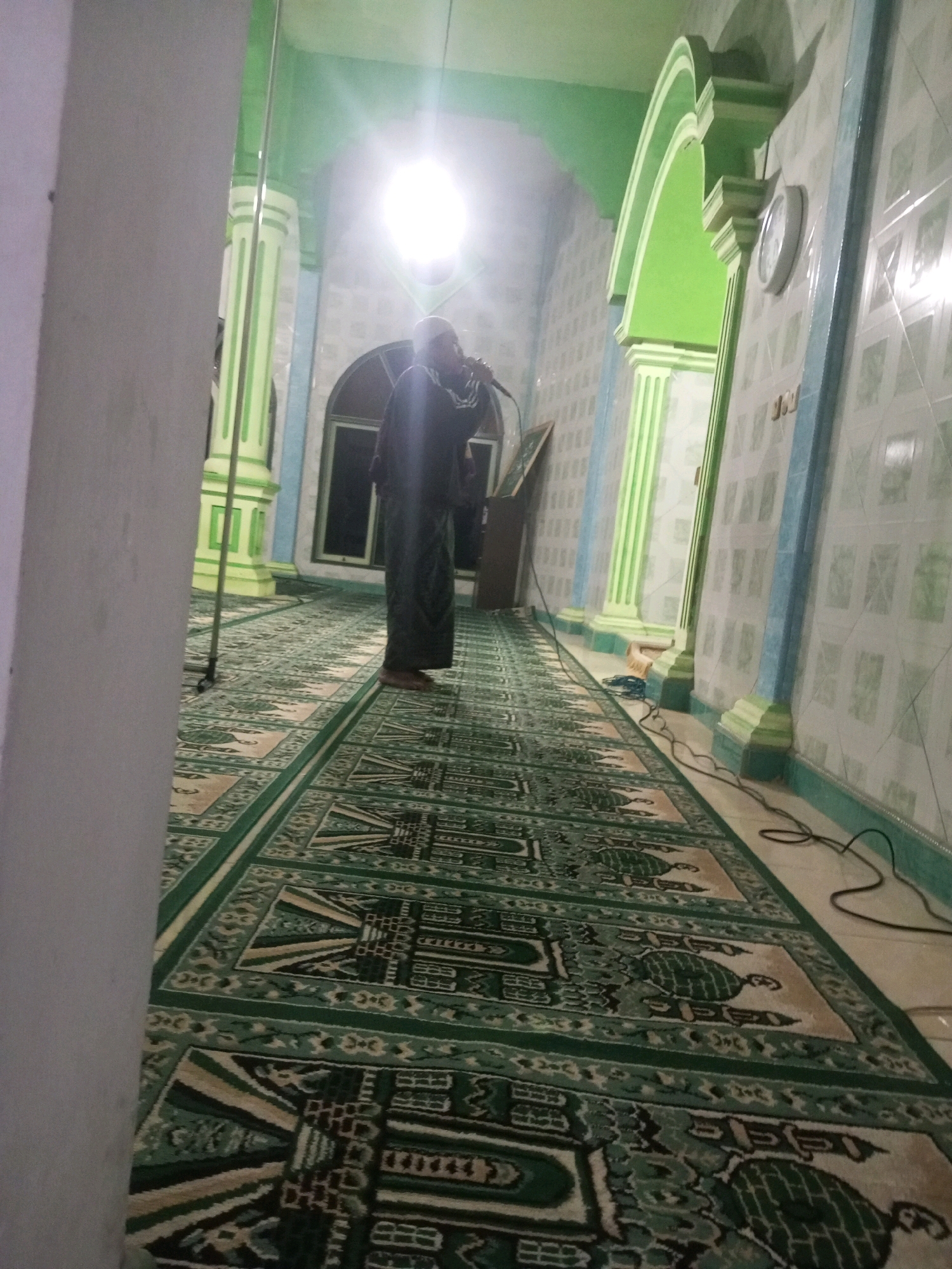 A person dressed in a dark robe and white head covering stands in a prayer hall, holding a microphone. The hall features green and white checkered tiled walls, a green carpet with a geometric pattern, and green arched pillars. A bright light emanates from the ceiling, creating a glare. Electrical cords are visible on the floor.