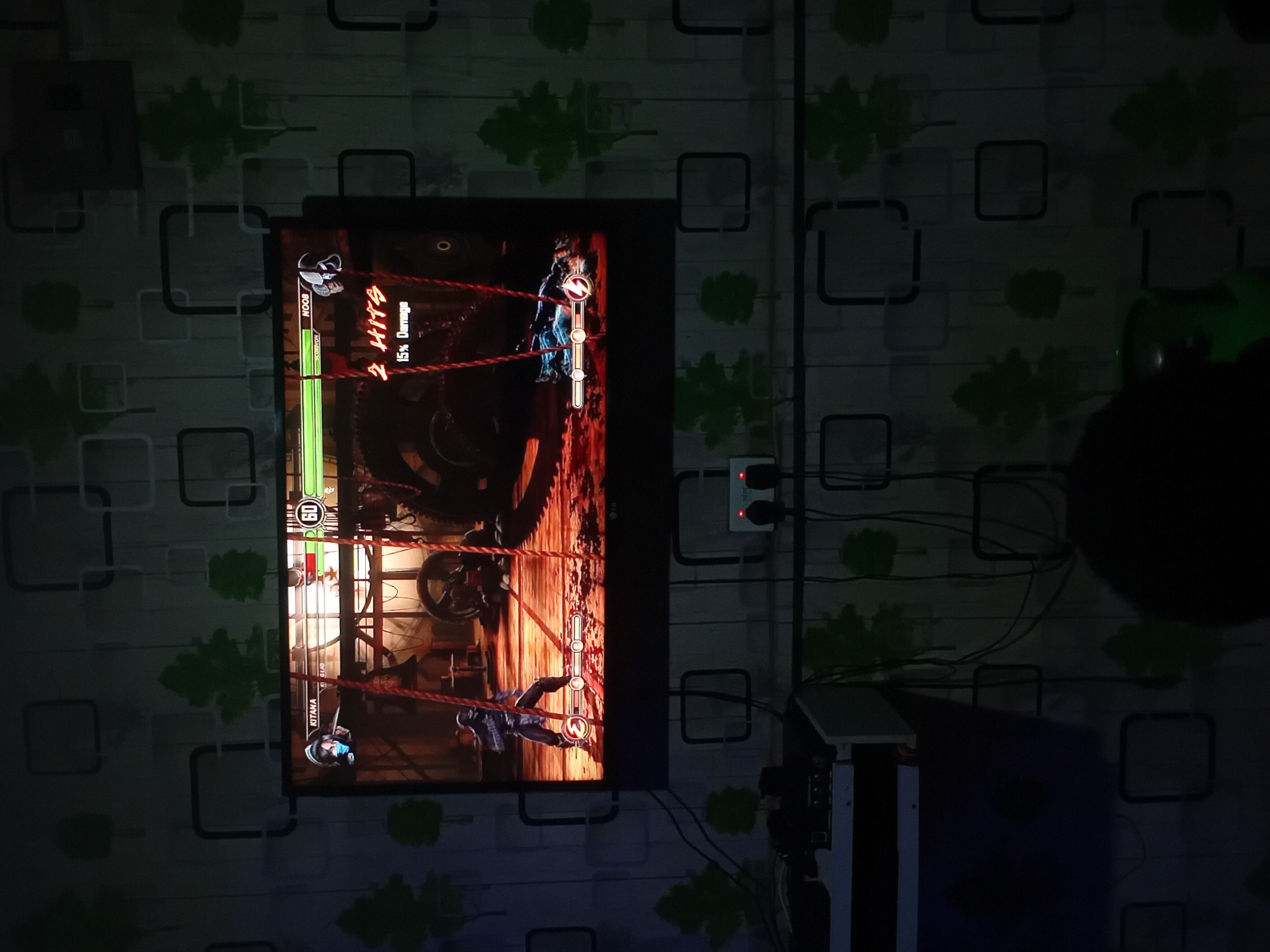 The image captures a dimly lit indoor room, likely at night, with the primary light source being a large TV screen displaying a video game. The wall behind the TV is covered in wallpaper featuring a pattern of light-colored squares and green leaf shapes.
The main subject is the TV screen, which shows a fighting game in progress. Two characters are visible: "NOOB" (Noob Saibot) at the top of the screen and "KITANA" at the bottom, indicating a match from the Mortal Kombat series. Noob's health bar is partially depleted, and a combo counter "2 HITS" and "15% Damage" is displayed next to him. Below Noob's name, the text "SCORPION" is visible, likely indicating the player's tag. Kitana's health bar is full. A timer displaying "60" is positioned at the top center of the screen, counting down. The game's background depicts an industrial or mechanical setting with large gears and red ropes.
To the right of the TV, a white power strip with red lights indicating active outlets is mounted on the wall, with various black wires running from it. Below the power strip and to the right of the TV, a dark, vertical object, possibly part of a computer tower or entertainment console, is partially visible. The overall scene suggests an immersive gaming environment in a residential setting. The monitor itself has a faint "LG" logo visible on its bottom bezel.