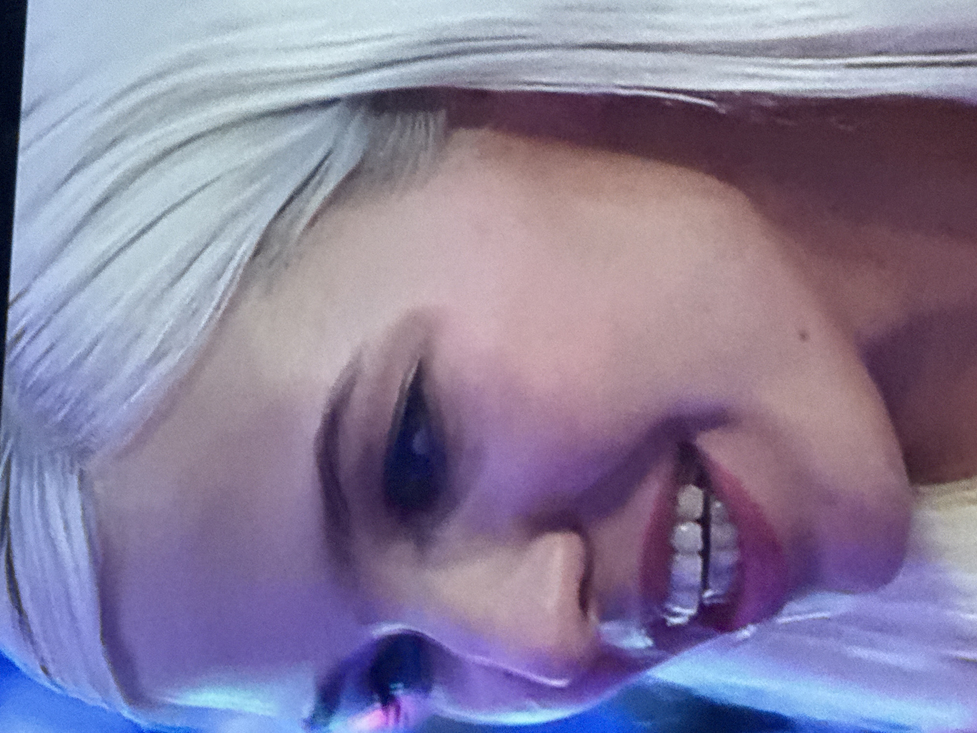 The image is a close-up shot of a fair-skinned individual, likely female, with bleached blonde hair. The person is smiling, revealing their teeth. They appear to have some dental work, possibly braces or retainers on their lower teeth. The lighting is somewhat blueish, possibly from a screen or other light source. The angle of the photo is slightly tilted. Location cannot be identified.
