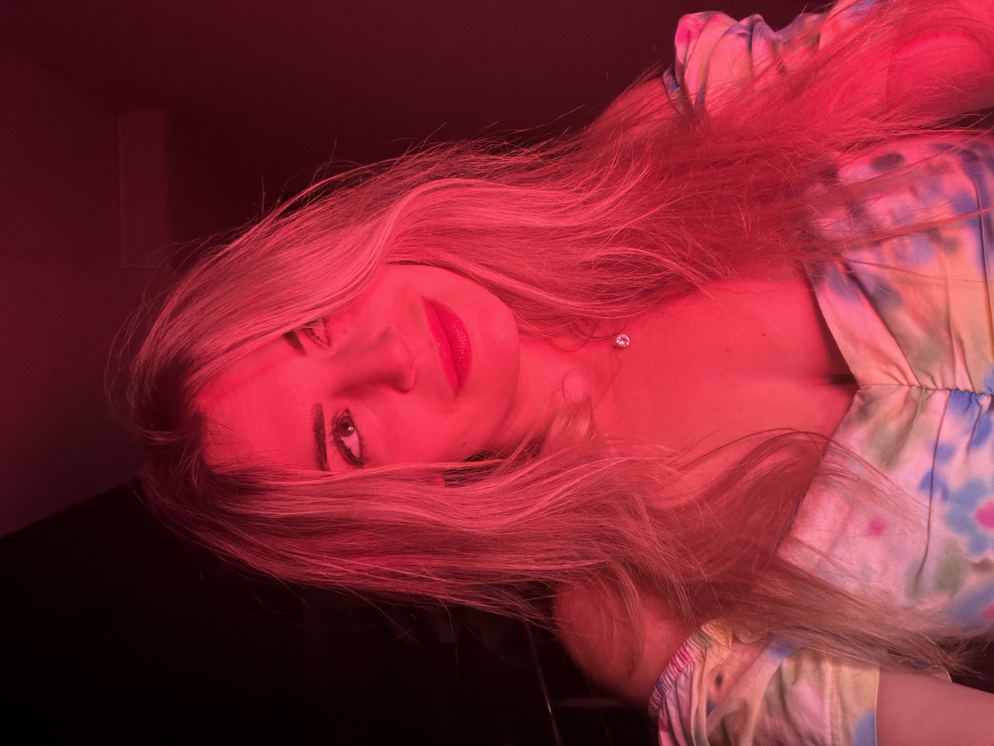 The image is a close-up portrait of a young woman illuminated by red lighting. She has long, blonde hair that falls around her shoulders. She is looking slightly upwards and to her left, with a subtle smile on her face. She is wearing a colorful off-the-shoulder top with a tie-dye pattern featuring pink, blue, and yellow hues. A delicate necklace with a small pendant is visible around her neck. The background is dark and out of focus, providing no specific environmental context or identifiable location within Budva, Montenegro. The red lighting casts a strong hue over the entire scene, obscuring finer details and creating a dramatic effect.