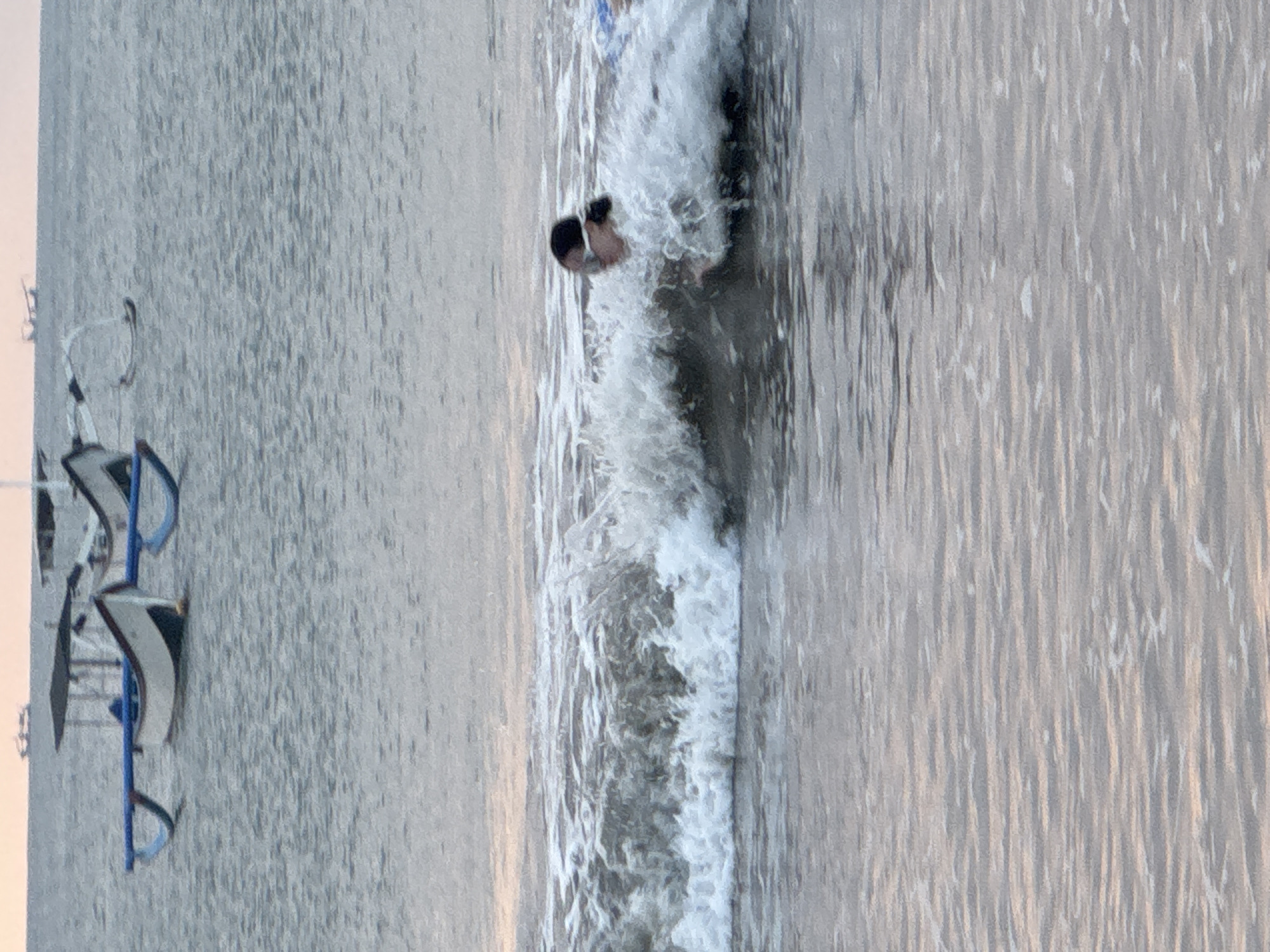 The image depicts a section of the ocean near the city of Kuta Selatan, Indonesia, at dusk. In the foreground, a wave is breaking, with a person wearing a snorkel mask partially submerged in the water, being enveloped by the foam. In the background, several traditional Balinese outrigger boats are visible on the calm water, with their distinctive hulls and outriggers. The sky above is a pale orange and grey, indicating the setting sun. The overall scene is peaceful and serene, with the natural elements of the sea and sky dominating the composition.