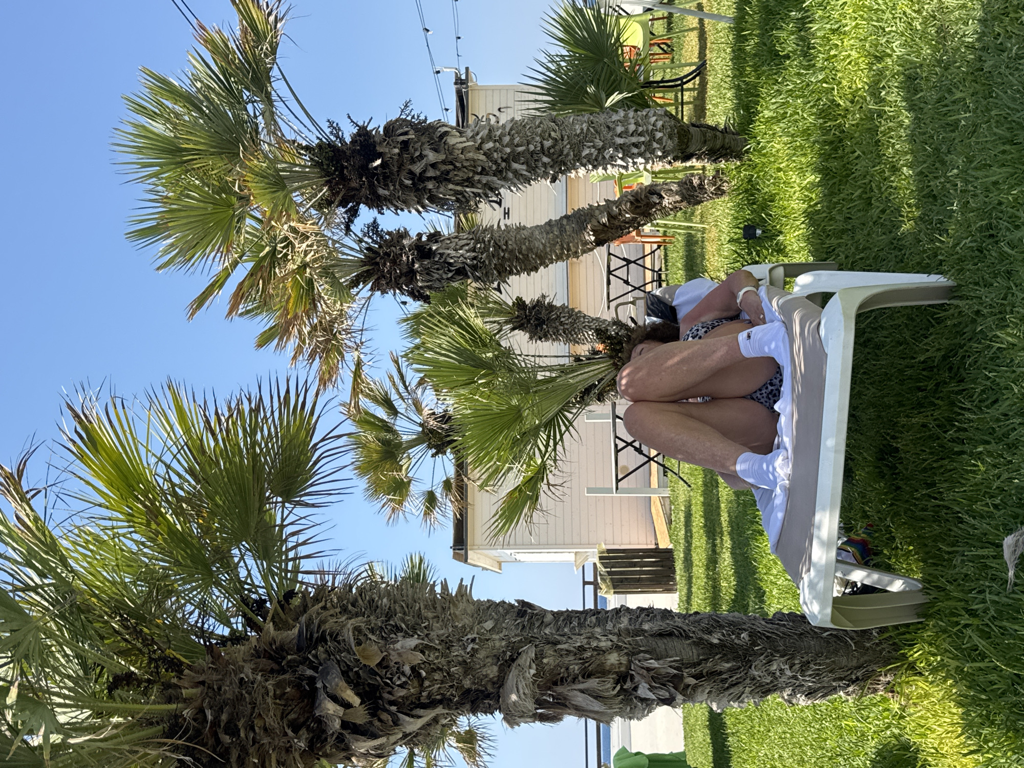 A person reclines on a white lounge chair on a grassy area. They are wearing a black and white patterned swimsuit and white socks. Their legs are crossed, with one foot resting on the other. Several palm trees surround the lounge chair, with their fronds casting shadows on the grass. In the background, a small white building with a dark roof is visible, along with some outdoor tables and chairs. The sky is clear and blue. The scene appears to be outdoors, likely in a warm climate, with the vegetation and building suggesting a coastal or resort location, possibly in Isola delle Femmine, Italy.