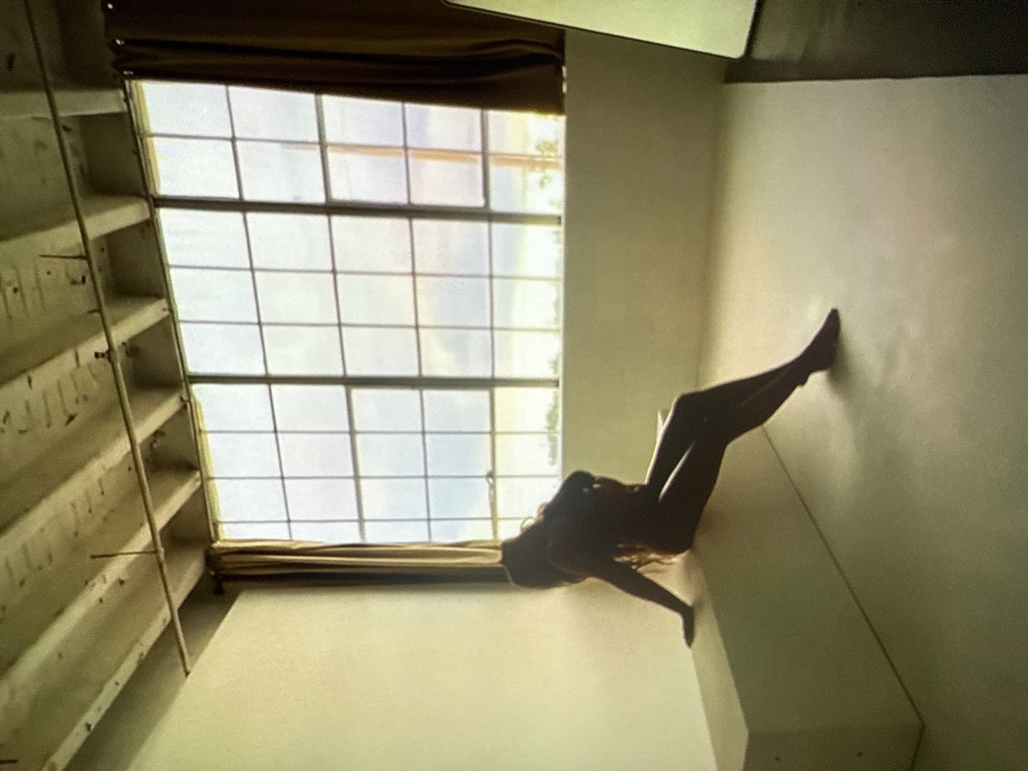 An indoor scene, likely a photographic studio or a loft, features a large, grid-paned window dominating the rear wall, serving as the primary light source. The room has light beige-colored walls and a matching floor, creating a minimalist backdrop. The ceiling displays exposed wooden joists and beams, beneath which a horizontal metal pipe or conduit runs, supported by a vertical rod from above. Dark brown curtains frame the large window on both sides. Through the window, an indistinct view of an overcast sky and distant structures is visible.
A single individual, appearing to be a female with long hair, is seated in profile on a low, rectangular, light-colored bench or platform positioned against the left wall. The person is rendered in a strong silhouette due to the backlighting from the window, revealing body contours consistent with nudity or minimal clothing. She leans back against the wall with her head tilted upwards and slightly backward. Her right arm extends behind her on the bench, while her left arm is bent, with her hand positioned near her chest area. Her legs are bent, with her feet resting on the floor in front of the bench. On the far right side of the image, a tall, narrow, light-colored rectangular panel or object, possibly a mirror or a reflective screen, is partially visible, standing vertically. The lighting creates a dramatic contrast, highlighting the silhouette of the person against the brighter window.
The image contains no specific visual cues or metadata to identify its geographical location as Varna, Bulgaria, or any particular city.