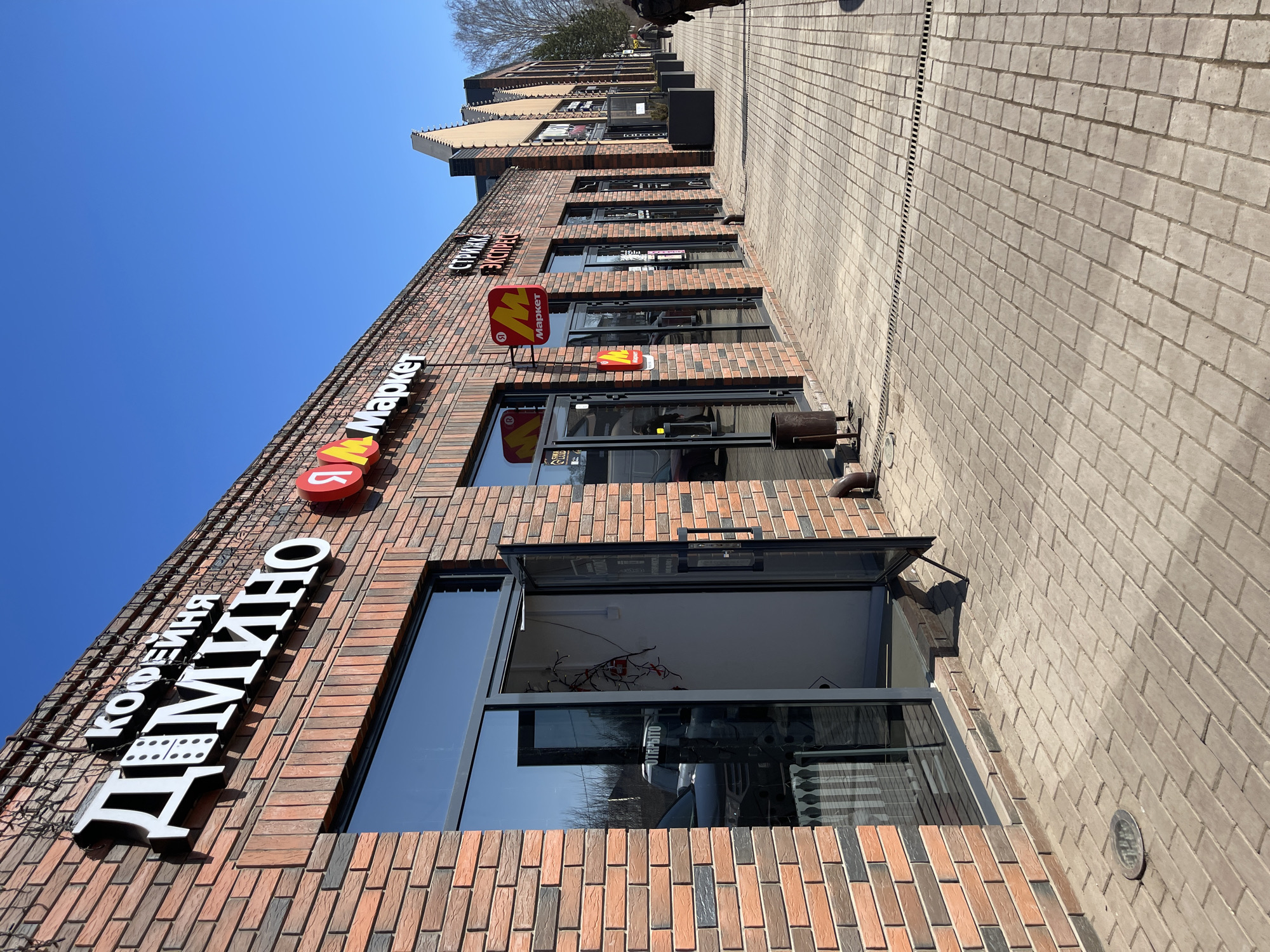 The image shows a commercial building in Osa, Russia, on a sunny day. The building is constructed of brick and features a row of storefronts. Signage is visible in Russian, including the word "Domino" (Дмитно) and "Market" (Маркет). The storefronts have glass doors and windows, some of which are open. A paved sidewalk runs alongside the building. There are no people visible in the image.