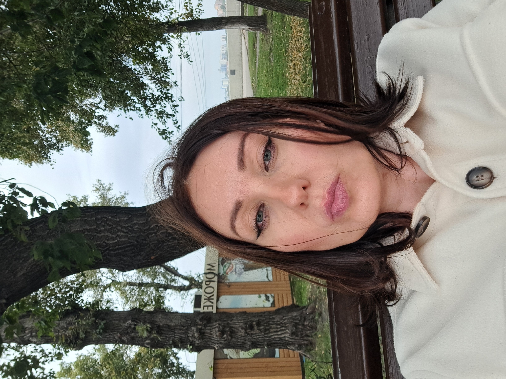 A woman with dark brown hair, fair skin, and blue eyes is taking a selfie while sitting on a wooden bench outdoors. She is wearing a cream-colored jacket with large brown buttons. Her lips are pursed and slightly parted, and her hair is blowing gently in the wind.
Behind her, two large trees with rough bark frame the scene. A wooden kiosk with the word "МОРОЖЕНОЕ" (ice cream) written in reversed Cyrillic is visible to the left of the woman. The kiosk has mirrored panels. To the right of the woman, there is a grassy area with fallen leaves and more trees. In the distance, a concrete barrier runs along what appears to be a waterfront, with a city skyline visible on the other side. The sky is overcast, suggesting a cool, cloudy day. The location is confirmed to be Irkutsk, Russia, given the Cyrillic signage.
