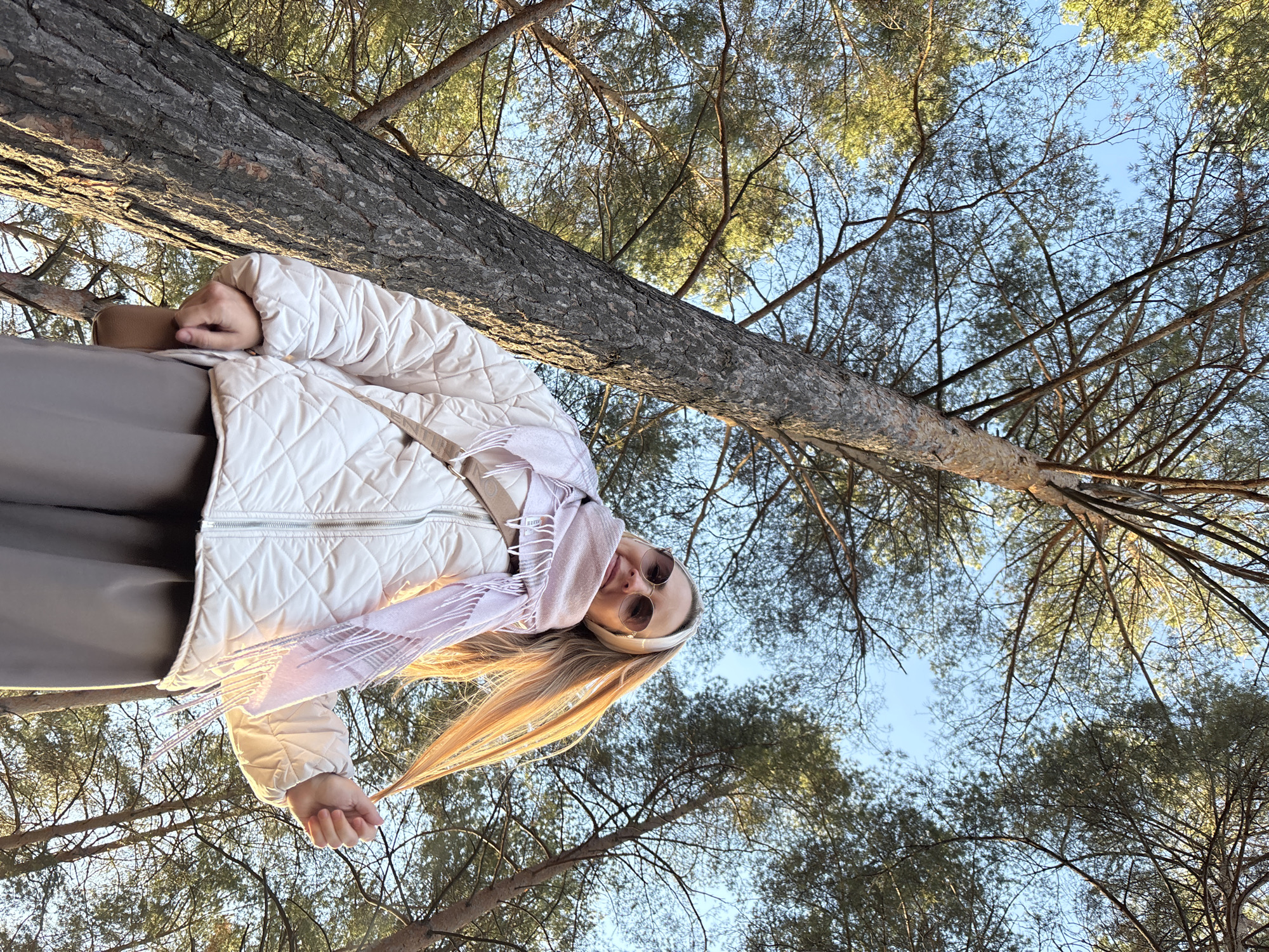 A person is captured in a low-angle shot with a tree trunk running diagonally across the frame. The person, with long blond hair, wears a white quilted jacket and a pale pink scarf. They are looking down at the camera with sunglasses on. The background consists of a dense canopy of trees against a pale blue sky. The location appears to be a forest. The image has natural sunlight and a slightly warm tone. No specific details indicate the location is in the city of Chaikovsky, Russia.