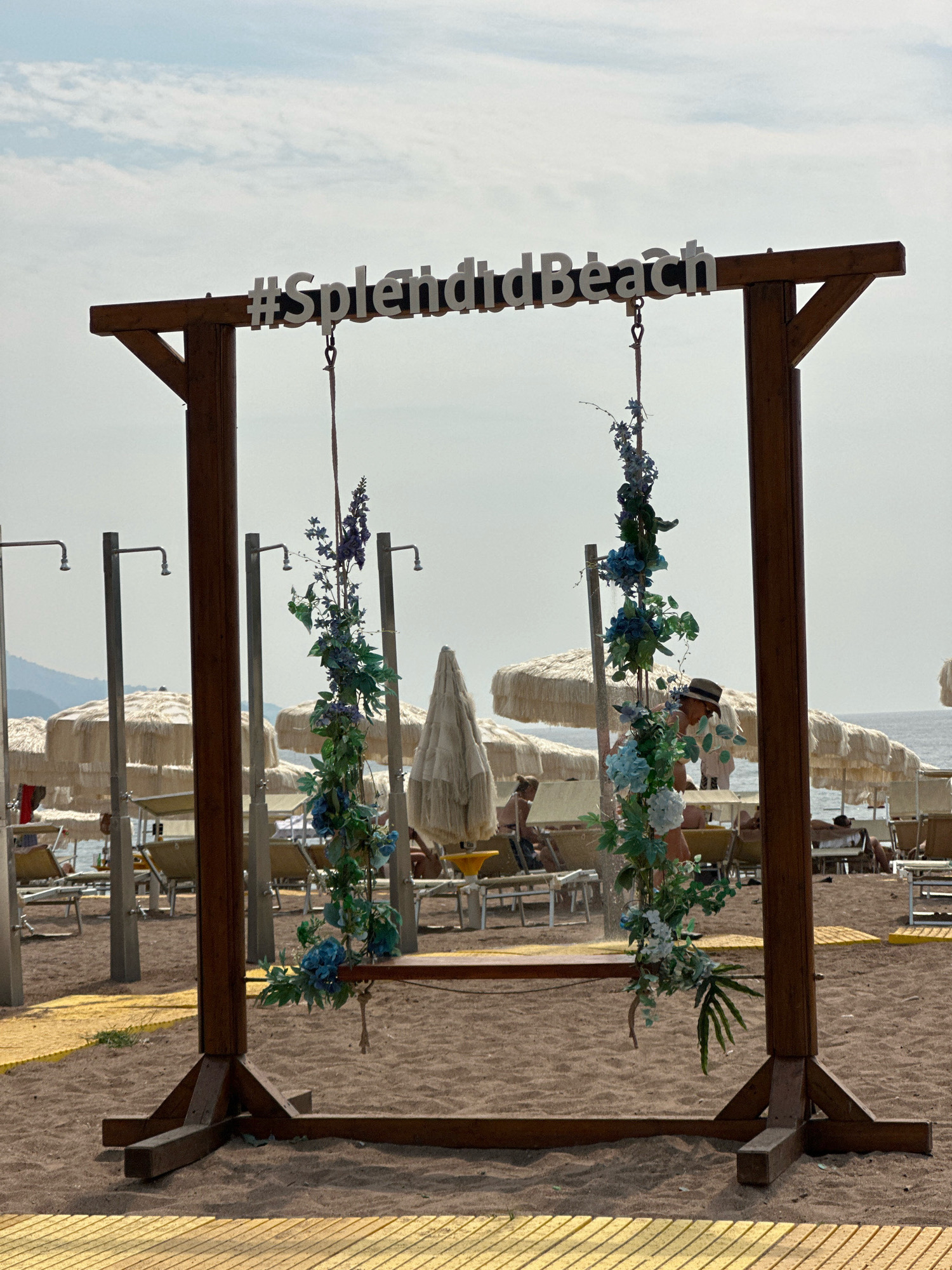 This is a full, eye-level, outdoor shot of a wooden swing set on a sandy beach. The swing set is adorned with artificial blue and white flowers and greenery. The word "#SplendidBeach" is spelled out in white, raised lettering across the top horizontal beam of the swing set. Two ropes hang from the top beam, supporting the wooden seat of the swing. In the background, there are numerous beach umbrellas, lounge chairs, and people relaxing on the beach. Several outdoor shower heads are visible on tall metal poles. The sea and a hazy mountainous coastline are visible in the distance. The scene appears to be set in the city of Boreti, Montenegro, given the context. The lighting suggests it is daytime, with overcast skies.