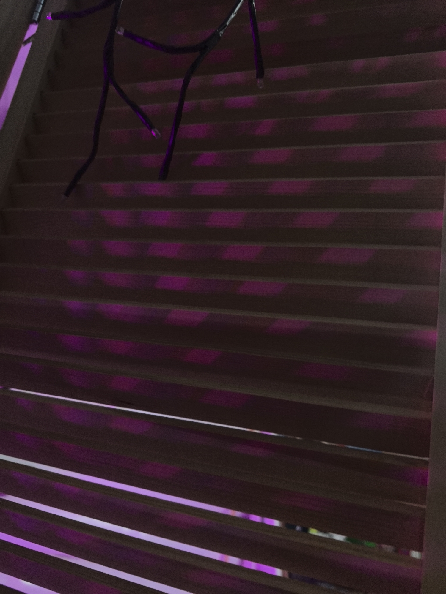 The image presents a dimly lit indoor scene featuring horizontal wooden slats, likely part of blinds, shutters, or a decorative screen. From the top left, a string of dark-colored decorative lights hangs downwards. Several individual light bulbs along this string emit a vivid purple or magenta glow.
The prominent feature of the image is the purple light, which casts distinctive, elongated rectangular reflections and illumination across the wooden slats. These vibrant purple patches are scattered across the entire surface, suggesting the light sources are positioned to shine through gaps in the slats or reflect off their surfaces. The slats themselves appear to be a natural wood tone, but their color is subdued by the low ambient light and intensified by the strong purple glow.
The overall atmosphere suggests a nighttime setting or a deliberately darkened room, where the decorative lights are the primary source of illumination. There are no people visible, nor any specific activity taking place. No text is present in the image. The provided crops offer closer views of the light string and the illuminated wooden surfaces, highlighting the purple reflections. The location context of Noviy, Russia, cannot be determined from the visual information within the image.