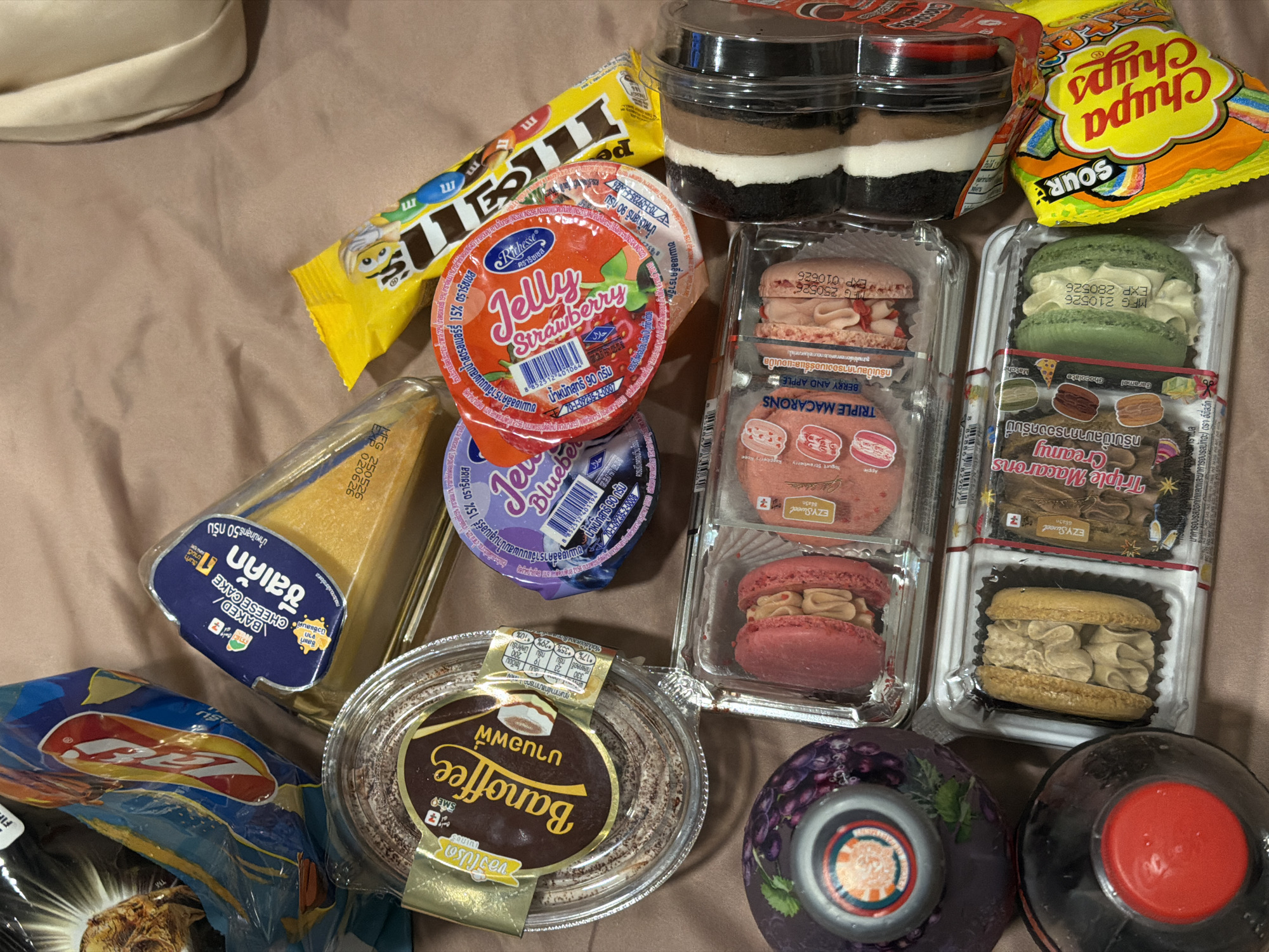 This image shows an assortment of food items placed on a brown surface. There's a yellow M&M's package, a red Jelly Strawberry container, and a blue Blueberry Jelly container. Also visible are two containers of what appears to be layered desserts and two packages of macarons from "EZY Sweet" with different flavors: one is labeled "Triple Macarons Creamy" with Caramel, Chocolate, and Matcha flavors, and the other is labeled "Triple Macarons Berry and Apple." Adjacent to the macarons is a package of "Sour Chupa Chups." Further to the left, there is a triangle-shaped container labeled "Baked Cheese Cake." There is a container labeled "Banoffee", and parts of bags of Lay's chips, and 2 covered round containers. There is no specific indication of the location being Kathu, Thailand, other than the text on the products suggesting a Thailand origin.