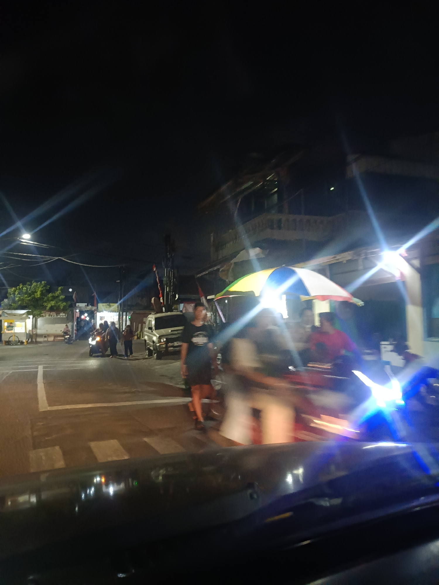 This is a nighttime, somewhat blurry photograph taken from inside a vehicle looking out at a street intersection. The scene appears to be in Surabaya, Indonesia. On the left side of the street, there's a food stall with a person attending it. A few people are gathered around a motorcycle in the middle of the intersection, and a silver SUV is parked to the right of the intersection. Another person, wearing a dark t-shirt and shorts, is walking in the foreground on the right side of the image. There are buildings and streetlights visible in the background, and colorful flags are displayed on poles. The overall impression is of a busy street scene at night, with a mix of pedestrians and vehicles.