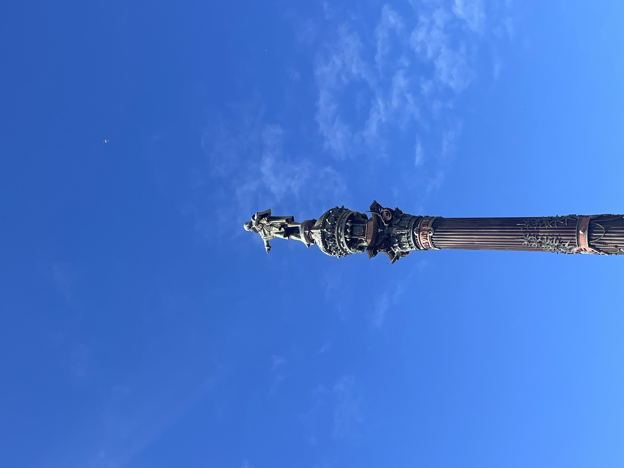 The image shows the Columbus Monument in Barcelona, Spain, viewed from below, with a blue sky background containing scattered clouds and a small flying object in the upper left corner. The monument's statue of Columbus and ornate pillar are prominently displayed. The absence of people in the frame emphasizes the monument's scale and design.