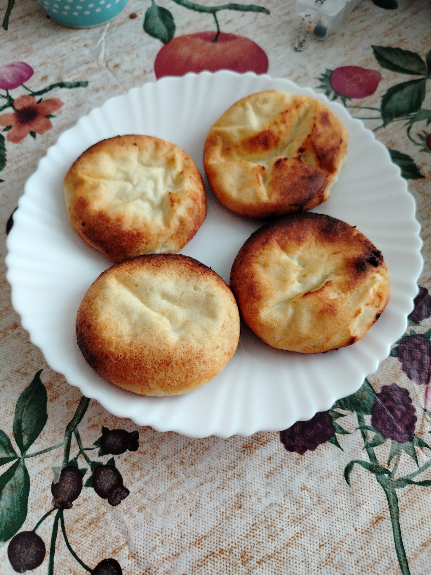 An overhead, close-up shot features four golden-brown baked goods arranged on a white plate with a scalloped edge. The items are round, with a slightly darker, crispy-looking exterior and a lighter, smoother center. They appear to be freshly baked, possibly a type of cheesecake or pancake (such as syrniki, given the context).
The plate rests on a tablecloth adorned with a colorful, patterned design. The pattern includes illustrations of red apples, red strawberries, dark purple blackberries, and various flowers (some purple, some orange, some pink) with green leaves and stems, all on a light, textured background.
In the upper left corner, a portion of a light blue mug with white polka dots is visible. In the upper right, a small, clear plastic container with a metallic charm or keychain attached is partially seen. The letters "RC" are faintly visible on the side of this container.
The lighting is bright and even, suggesting a daytime setting, possibly indoors. The overall scene conveys a homely and inviting atmosphere, likely depicting a meal or snack prepared in Chaikovsky, Russia. No specific activity or emotions are directly conveyed, but the presentation of the food suggests it is ready to be eaten.