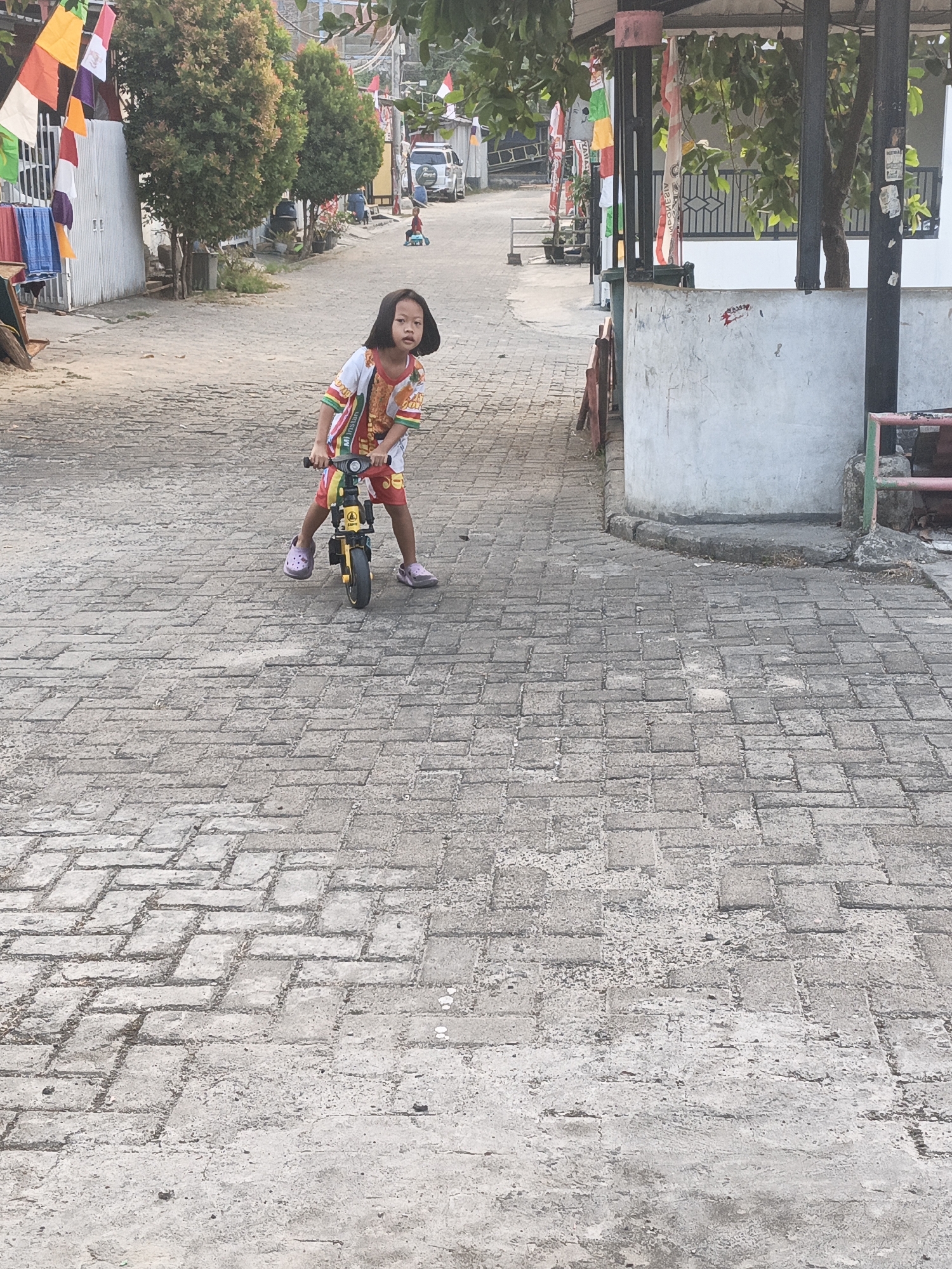 A young girl with dark hair is on a yellow balance bike in a paved alleyway in Cilegon, Indonesia. She is wearing a white shirt with colorful patterns and red shorts. Her feet are on the ground, and she appears to be in motion. The alleyway is paved with grey brick. In the background, there are houses, trees, and some flags displayed. A white car is parked further down the alley.