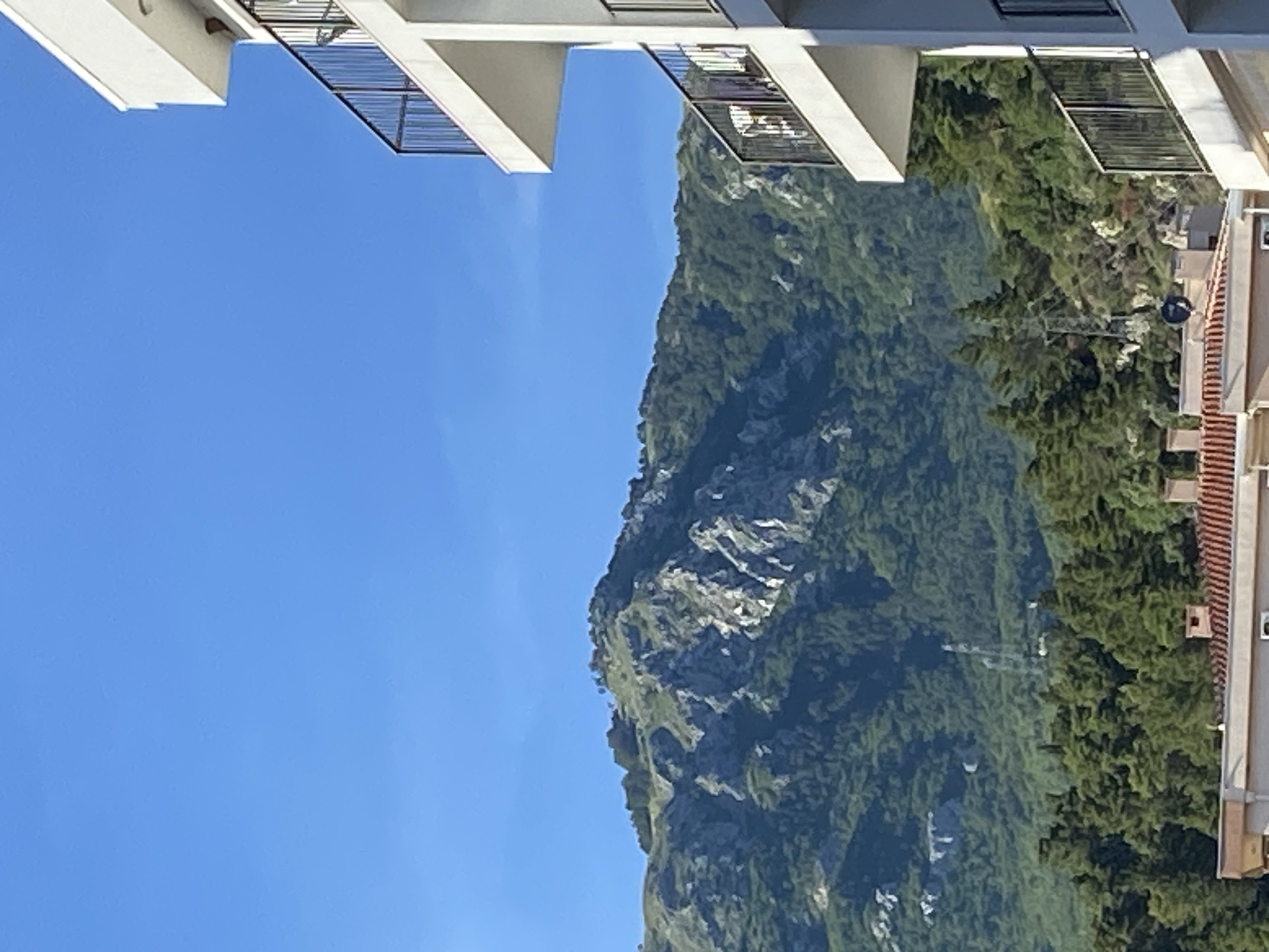 The image depicts a bright, clear day in a coastal mountainous region, identified as the city of Budva, Montenegro, based on its characteristic landscape. The upper half of the frame is dominated by a clear, vivid blue sky with no discernible clouds. Below the sky, a large, rugged mountain occupies the mid-ground, featuring significant exposed grey rock formations interspersed with dense, dark green foliage, primarily coniferous trees and shrubs. Near the center-right slope of the mountain, a tall, slender communication tower or similar antenna structure is visible, partially obscured by trees.
In the foreground, two distinct building structures are visible. On the right side, a modern, multi-story white building with flat roofs and pronounced balconies extends from the top to the bottom edge. Each balcony features black metal railings. On the second visible balcony from the top, a white towel or piece of fabric is draped over the railing. Further down, another balcony appears to contain dark-colored outdoor furniture. On the bottom-left, the terracotta-tiled roof and a single chimney of another building are partially visible, situated below a dense cluster of green trees that partially obscure the base of the mountain. No individuals or specific actions are discernible in the scene.