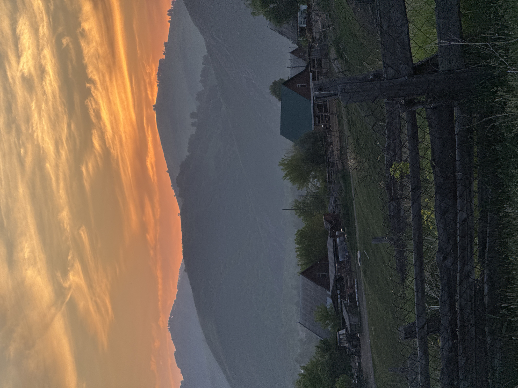 The image depicts a rural landscape at dusk, with rolling hills silhouetted against an orange and yellow sky. In the foreground, a wooden fence made of rough-hewn logs and wire mesh separates a grassy area from a cluster of farm buildings and agricultural equipment. The buildings appear to be simple wooden structures, some with dark roofs and walls, and one with a green roof. A tractor and other machinery are visible near the buildings. The environment is serene and quiet, characteristic of a rural village in the Altai Krai region, possibly near Soloneshenoe, Russia, given the mountainous terrain and rural architecture. There are no people or animals visible in the frame.