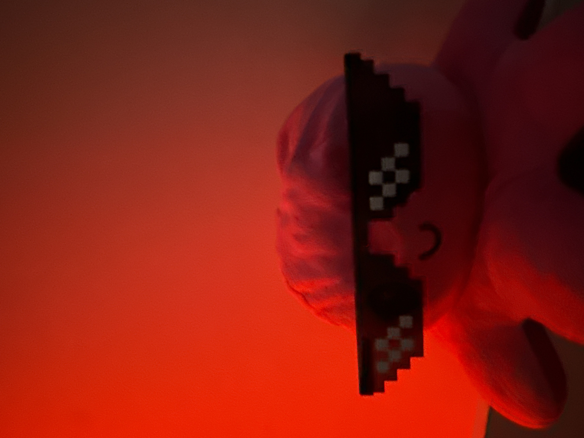 A stuffed octopus toy wearing pixelated sunglasses is positioned against a red background. The toy has a pink hue and a simple curved mouth. The background is a gradient of red to a lighter orange hue. The image is taken from a low angle, with the toy occupying the bottom half of the frame. There are no discernible features of Varna, Bulgaria, in this image.