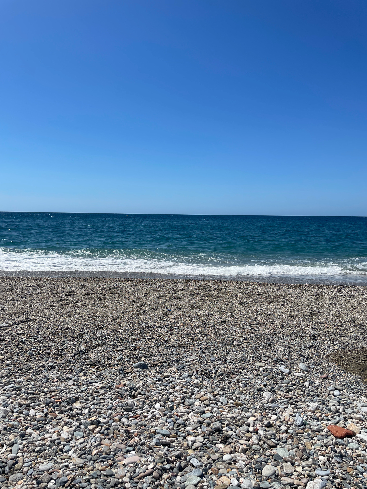The image shows a pebble beach with the Mediterranean Sea and a clear blue sky. The beach is covered in smooth, grey and white pebbles of various sizes. The sea is a deep blue color, with small waves breaking on the shore, creating white foam. The sky is a vibrant, cloudless blue. The location is the beach of Torrenueva Costa, Spain. There are no people or objects in the frame, only the natural elements of the beach, sea, and sky.