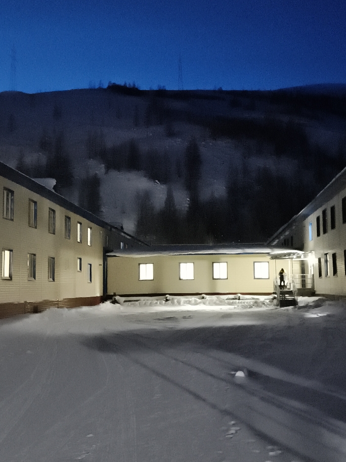 This image depicts a snow-covered outdoor courtyard in the city of Im. Gastello, Russia, at twilight or night. Two multi-story buildings, light-colored with darker lower sections, form an L-shape around the courtyard. Numerous rectangular windows are visible across both structures; many are illuminated, casting bright light onto the snow.
The courtyard ground is uniformly covered in snow, showing areas of disturbance and potential tracks. On the right side of the image, near the corner where the two buildings meet, a single individual stands on a short set of outdoor steps with railings. This person is facing generally towards the viewer, dressed in a heavy, possibly yellow or orange, winter jacket with a hood. An exterior light fixture illuminates the area around the steps.
In the background, behind the buildings, a large, dark, snow-covered mountain or hill range extends across the horizon, topped by a deep blue sky. Sparse dark trees are discernible on the lower slopes of the distant terrain.