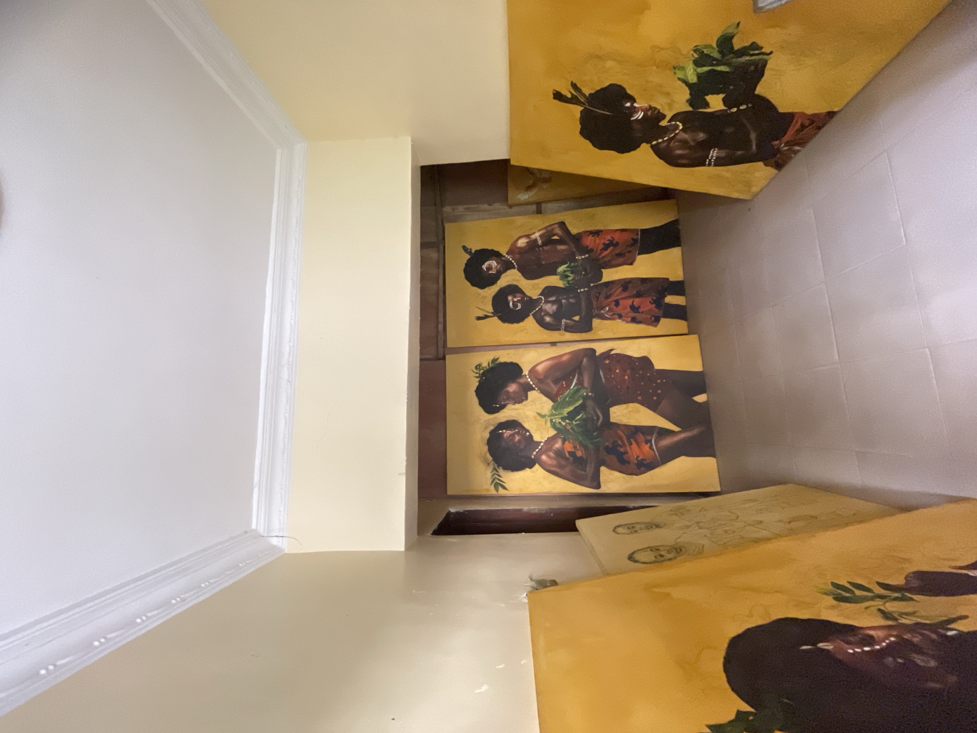 The image shows a collection of framed paintings leaning against a wall. The paintings are styled in an artistic, vibrant format and placed on a wall. The specific location in Ado, Nigeria, cannot be determined from the image. The interior shows white walls.