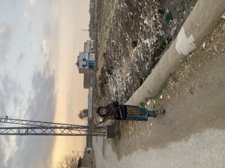 A young girl, approximately 5-8 years old, stands on a dirt path adjacent to a paved road in Quetta City Tehsil, Pakistan. She is dressed in a dark top, a patterned teal skirt with gold embellishments, and a patterned headscarf, looking directly at the camera with her arms crossed. To her left, a tall metal lattice utility pole rises. Parallel to the dirt path and to her right, a concrete-lined channel contains stagnant water and refuse. Beyond this channel, a large open field is extensively covered with accumulated solid waste, including plastic and other debris. In the mid-ground, a light-colored two-story building with blue trim and a small dome on its roof is visible behind a low wall bearing script. Other less distinct buildings are in the background. A male figure is seen walking away on the paved road in the far left background. The sky above is overcast with an orange-yellow hue near the horizon, suggesting late afternoon or dusk.