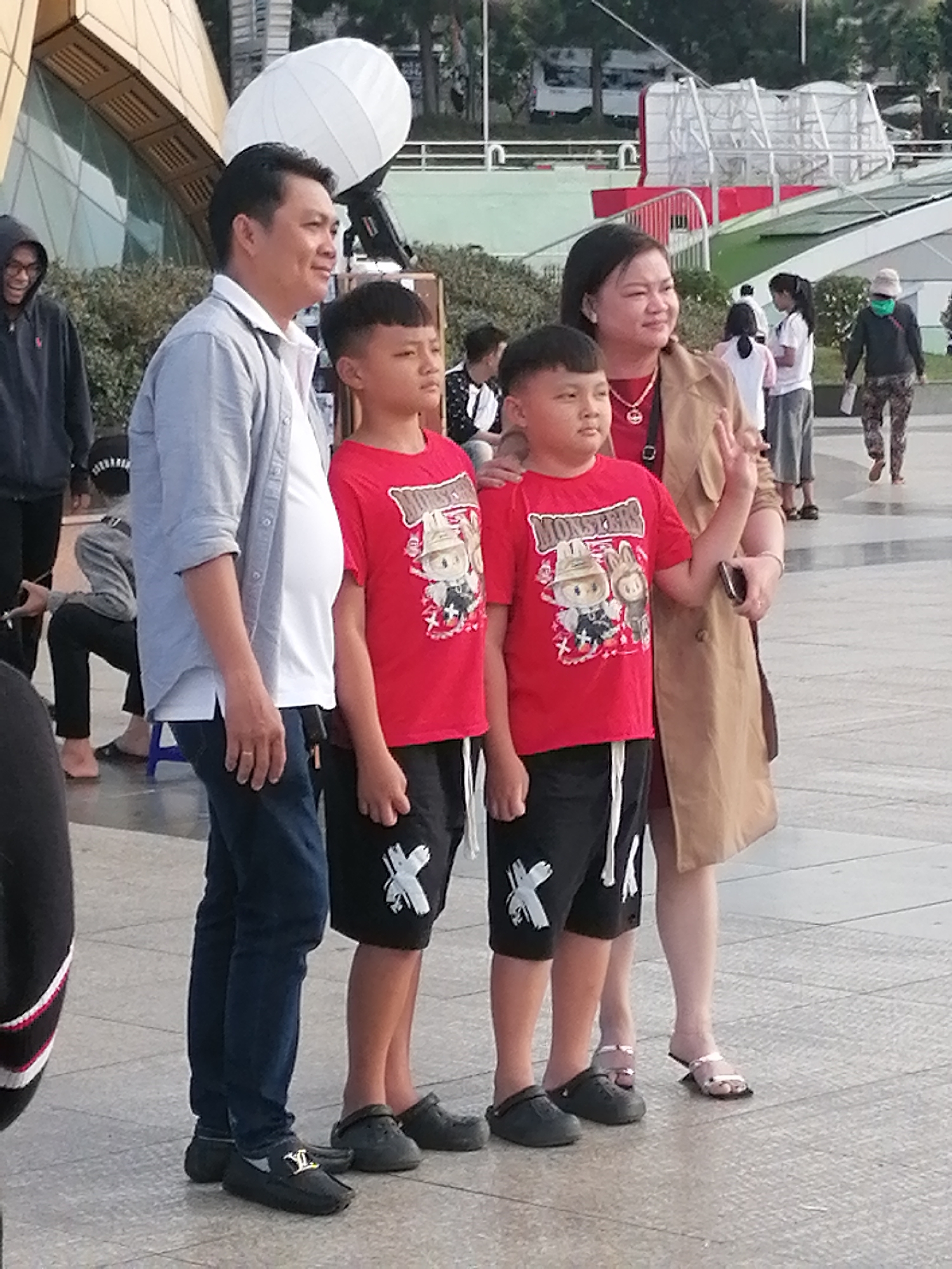 The image depicts a family standing outdoors in Da Lat, Vietnam. In the foreground, a man in a light blue shirt and dark jeans is standing next to his son, who is wearing a red t-shirt with a cartoon character and black shorts. The man's wife, in a tan coat and red shirt, stands next to another son, who is also wearing a red t-shirt and black shorts. The family appears to be posing for a photo, with the mother making a peace sign. In the background, other people are visible, including a person wearing a black hoodie, another person in a black jacket and camouflage pants, and a woman in a white shirt and gray skirt. The environment seems to be a public space with a paved ground and some greenery. A large white spherical object, possibly a balloon or lantern, is visible in the upper left corner of the image, with a structure resembling a telescope mount behind it. The architecture in the background suggests a modern building with glass panels. The overall scene appears to be a casual outing in a tourist area of Da Lat.