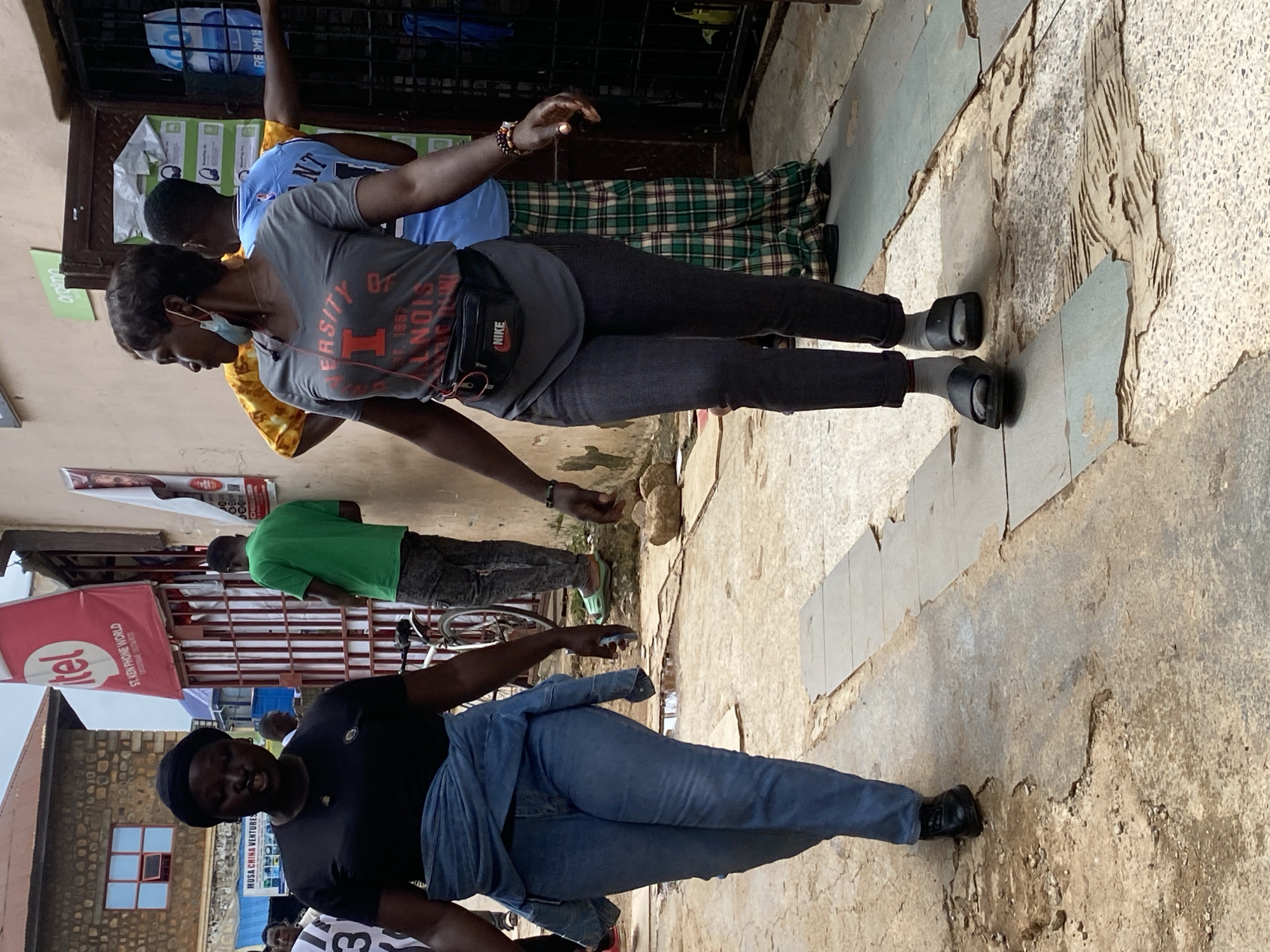 In an outdoor marketplace setting, two women are walking towards the viewer. The woman on the left, wearing a black t-shirt, blue jeans, and a black headscarf, is speaking animatedly. She has a denim jacket tied around her waist. The woman on the right is wearing a gray t-shirt with "UNIVERSITY OF ILLINOIS" printed on it in red, dark gray pants, and black slides. She has a black fanny pack with the Nike logo around her waist and is wearing earbuds. Behind them, several other individuals are visible, including a man in a green t-shirt and another in a blue basketball jersey with the name "MCINTOSH" and the number "12" on it. A person in a plaid green and white garment is also present. The ground is paved with concrete tiles, some of which are broken and weathered, creating a rough texture. Buildings with stone and painted walls line the sides of the narrow street, with shop entrances and various signs visible, including one for "TEL" and another for "MUSA CHINA VENTURES." The overall environment suggests a bustling urban marketplace.