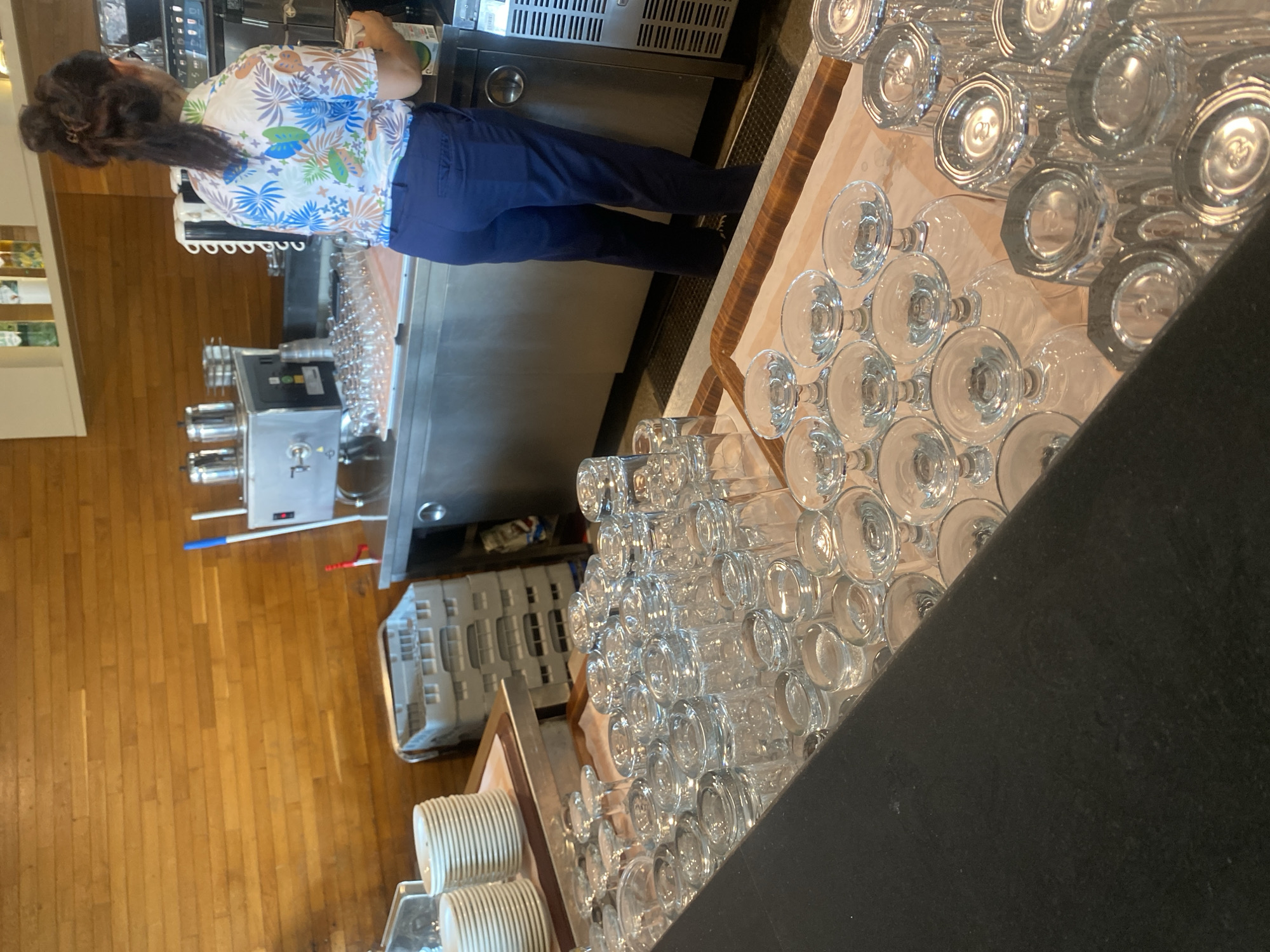 The image shows a woman in a colorful floral shirt and blue pants standing behind a stainless steel counter in what appears to be a bar or restaurant. She is holding a white carton and looking down. In front of the counter, there are many stacks of clean glassware, including tumblers and stemmed glasses, arranged on trays. Behind the woman, there is a stainless steel coffee or water machine, and to the left, stacks of white plates are visible. The wall behind the counter is made of wooden planks. There are no discernible landmarks or clues within the image to suggest a specific location in Aksu, Turkey.