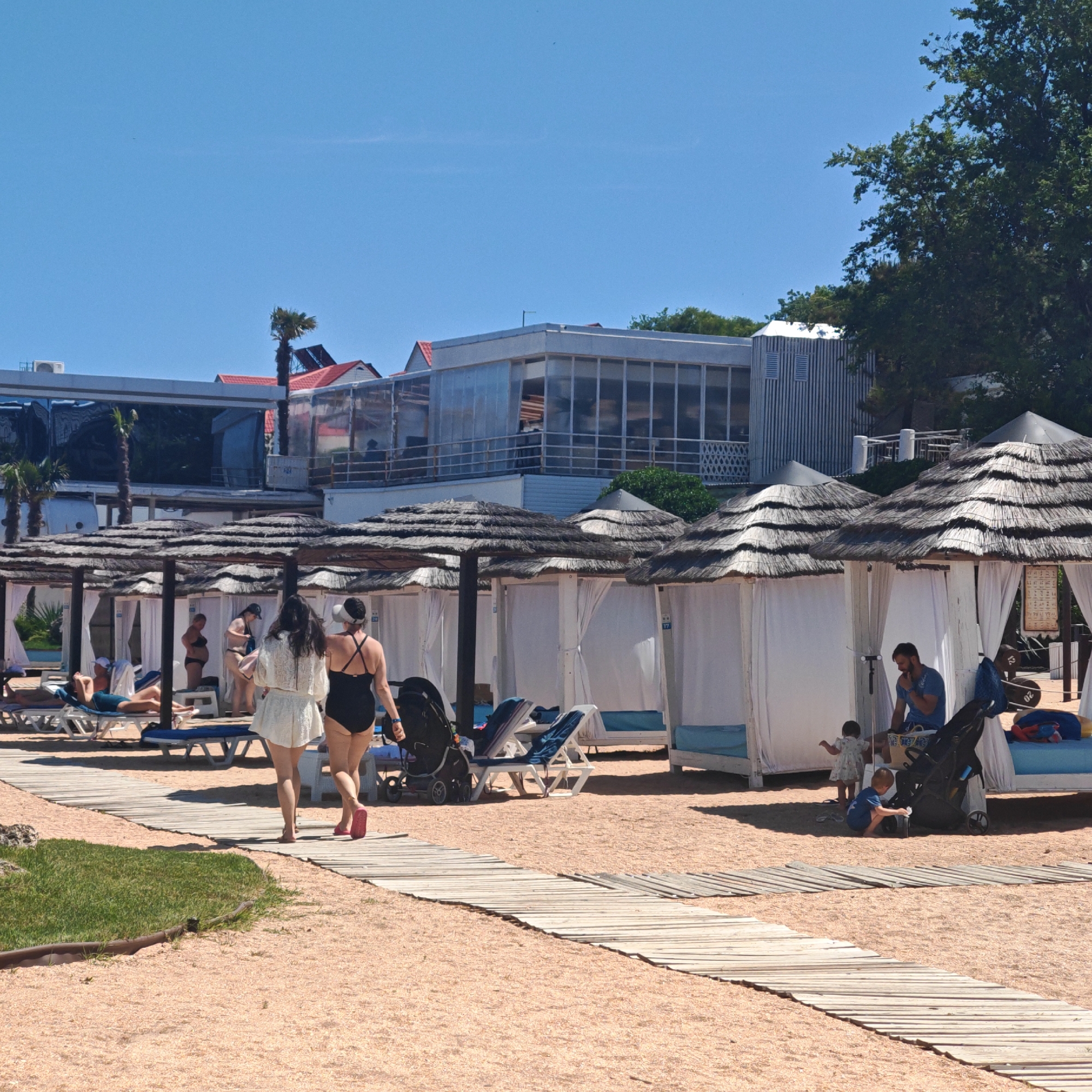This image captures a sunny beach scene at an unspecified resort location in Unknown, Unknown. The foreground features light brown sand with a segment of green grass on the left and a wooden boardwalk extending from the lower left towards the upper right.
In the mid-ground, numerous open-sided beach cabanas or pergolas with thatched roofs and white fabric curtains line the sandy area. Many contain blue and white lounge chairs. People are engaged in various leisure activities:
* Two women walk away from the camera on the boardwalk; one wears a white two-piece outfit, and the other, in a black one-piece swimsuit and white visor, pushes a black baby stroller.
* To the left, under cabanas, a woman in a dark swimsuit reclines on a lounge chair, while another woman in a black bikini stands within a cabana. Other individuals are seated or standing further back.
* To the right, under a cabana, a bearded man in a blue t-shirt sits on a lounge chair. Nearby, two small children are present: one child in a light dress stands, and another in a blue shirt kneels on the sand next to a black stroller.
The background reveals multi-story buildings with extensive glass windows and some red-tiled roofs, indicative of a hotel or resort complex. Green trees, including palm trees, are visible between and behind the structures. The sky is clear and bright blue. The scene is illuminated by direct sunlight, casting shadows.
