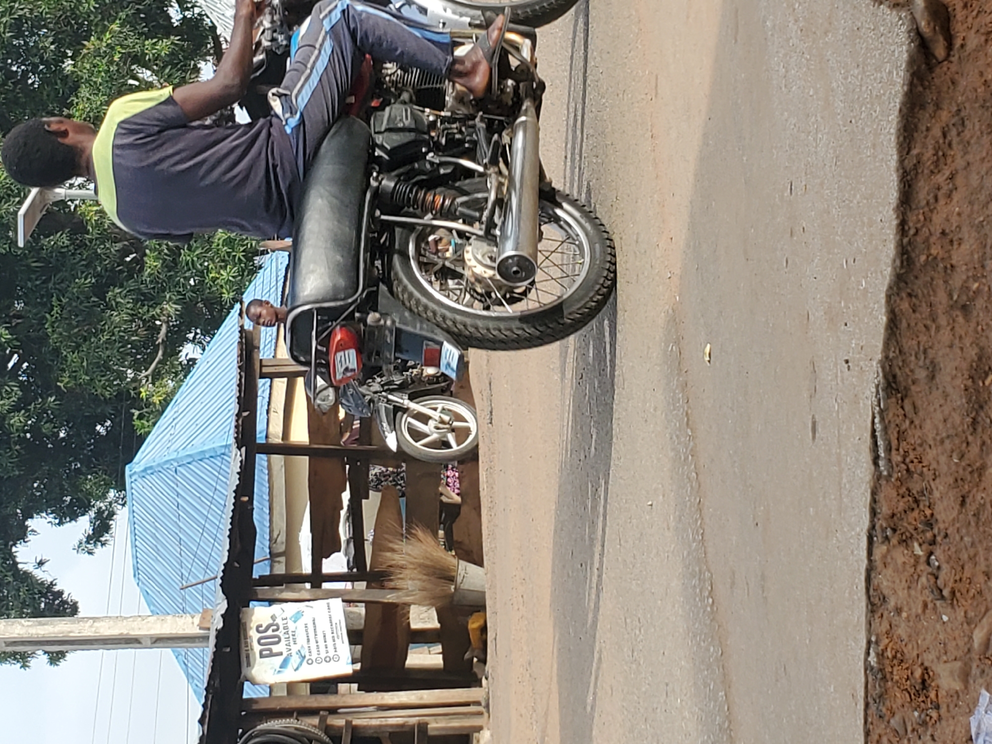 The image captures a vibrant roadside scene, likely in Mararaba, Nigeria, under bright daylight. In the foreground, a person is seated on a black motorcycle, facing away from the viewer. They are dressed in a dark blue short-sleeved top with light green shoulders and dark track pants with light blue stripes, with bare feet resting on the motorcycle's footpegs. The motorcycle itself appears to be a standard model, featuring a prominent silver exhaust pipe.
To the right of the foreground motorcycle, a paved road or a well-trodden path transitions into a dirt shoulder. In the middle ground, on the left, lush green trees provide a natural backdrop. To the right, a roadside stall or small shop is visible, characterized by its light blue corrugated metal roof. A person's head can be seen peeking out from behind the foreground motorcycle, near this stall, suggesting another individual is present.
The stall has various items, including a traditional broom and another motorcycle parked further back. A white sign affixed to the stall clearly advertises "POS AVAILABLE HERE." Below this, a list of services is visible, including "CASH WITHDRAWAL," "FUND TRANSFER," "BILL PAYMENT," "AIRTIME & DATA," "ACCOUNT OPENING," "BVN ENROLLMENT," and "NIN REGISTRATION." The bright sunlight indicates it is daytime, casting clear shadows. The overall setting depicts a common scene of daily commerce and transportation in a bustling local environment.