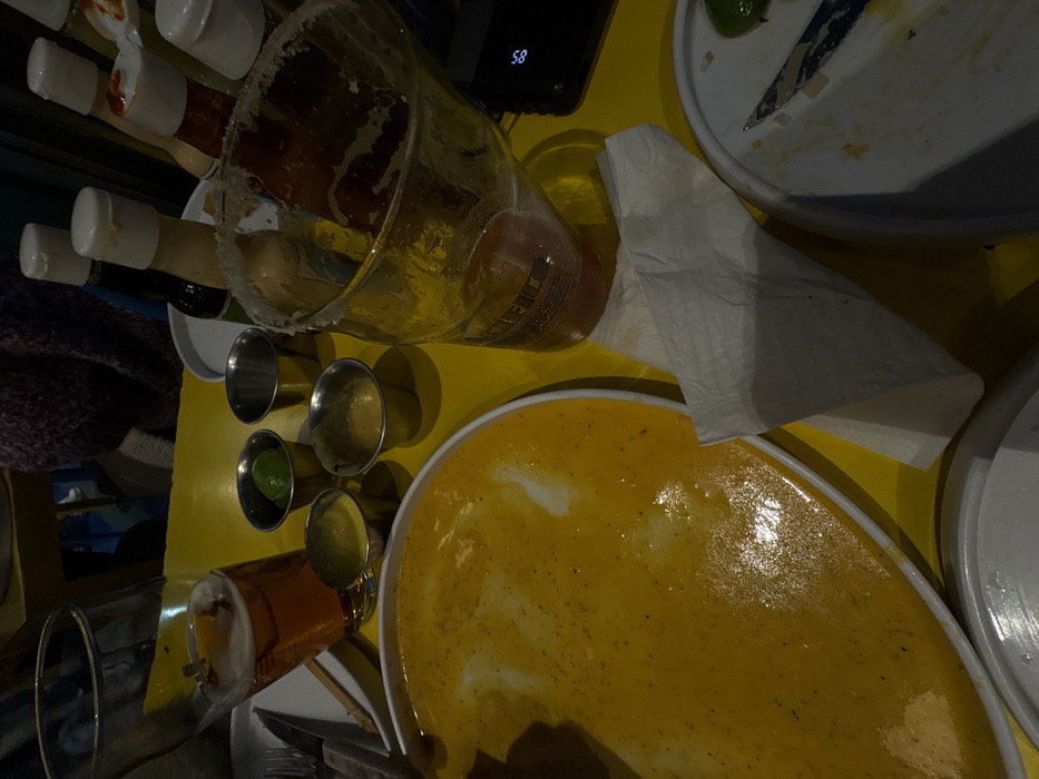 An overhead shot captures a messy, yellow table setting after a meal. A large, mostly empty plate of yellow food dominates the scene, accompanied by condiment bottles and small metal ramekins. A salt-rimmed glass lies on its side. A crumpled white napkin is partially obscuring another plate and a cell phone. A second glass sits near silverware on the table edge.