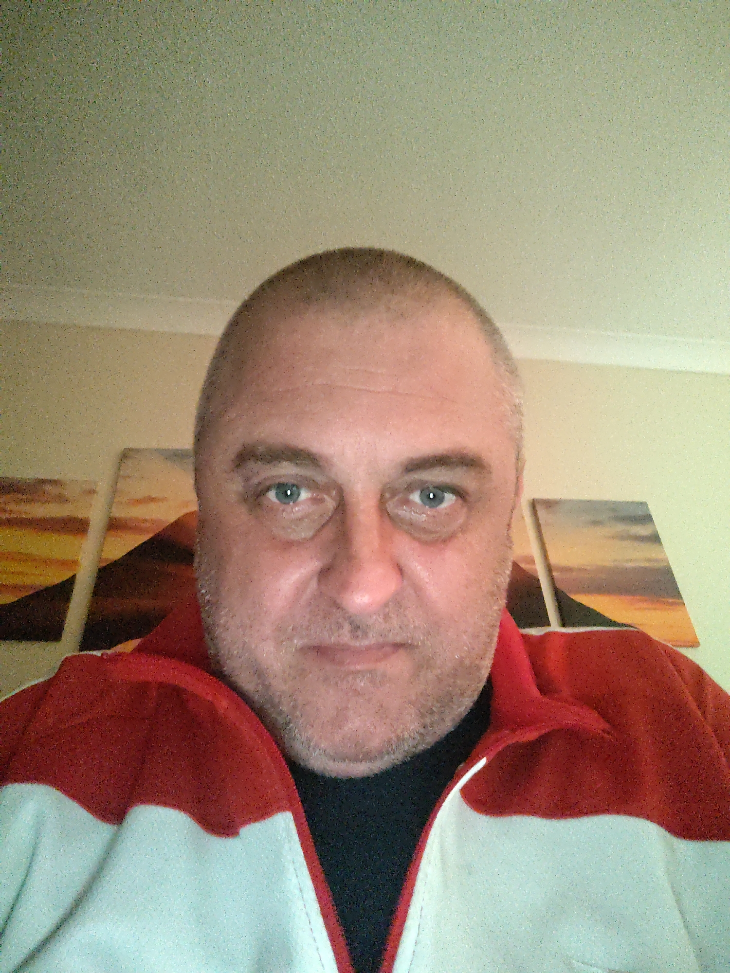 A man with light blue eyes and short, buzzed hair is captured in a close-up, head-and-shoulders selfie, looking directly at the camera. He has a neutral to slightly pleasant expression, with visible stubble on his chin and cheeks. He is wearing a red and white collared jacket or fleece over a black t-shirt or shirt.
The setting appears to be indoors, likely a room in a residence in Huntingdonshire, United Kingdom. The background features a light-colored wall, possibly cream or beige, with the ceiling visible at the top of the frame. Behind the man's left shoulder, two framed pictures or canvases depicting a warm-toned landscape, possibly a sunset or sunrise over a mountain or hill, are partially visible. A third similar picture is faintly seen behind his right shoulder. The lighting suggests an indoor environment. There is no visible text in the image.