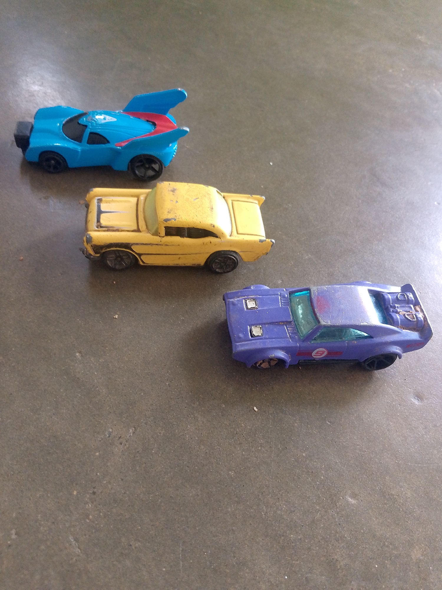 Three toy cars are placed on a dark, textured surface. The car furthest back is bright blue with red accents on its spoiler. In the middle, a yellow toy car with a worn finish. The car closest to the viewer is purple with some red accents and appears to be missing a wheel. The cars are arranged diagonally across the frame. The location of La Tagua, Venezuela is not depicted or suggested in the image.