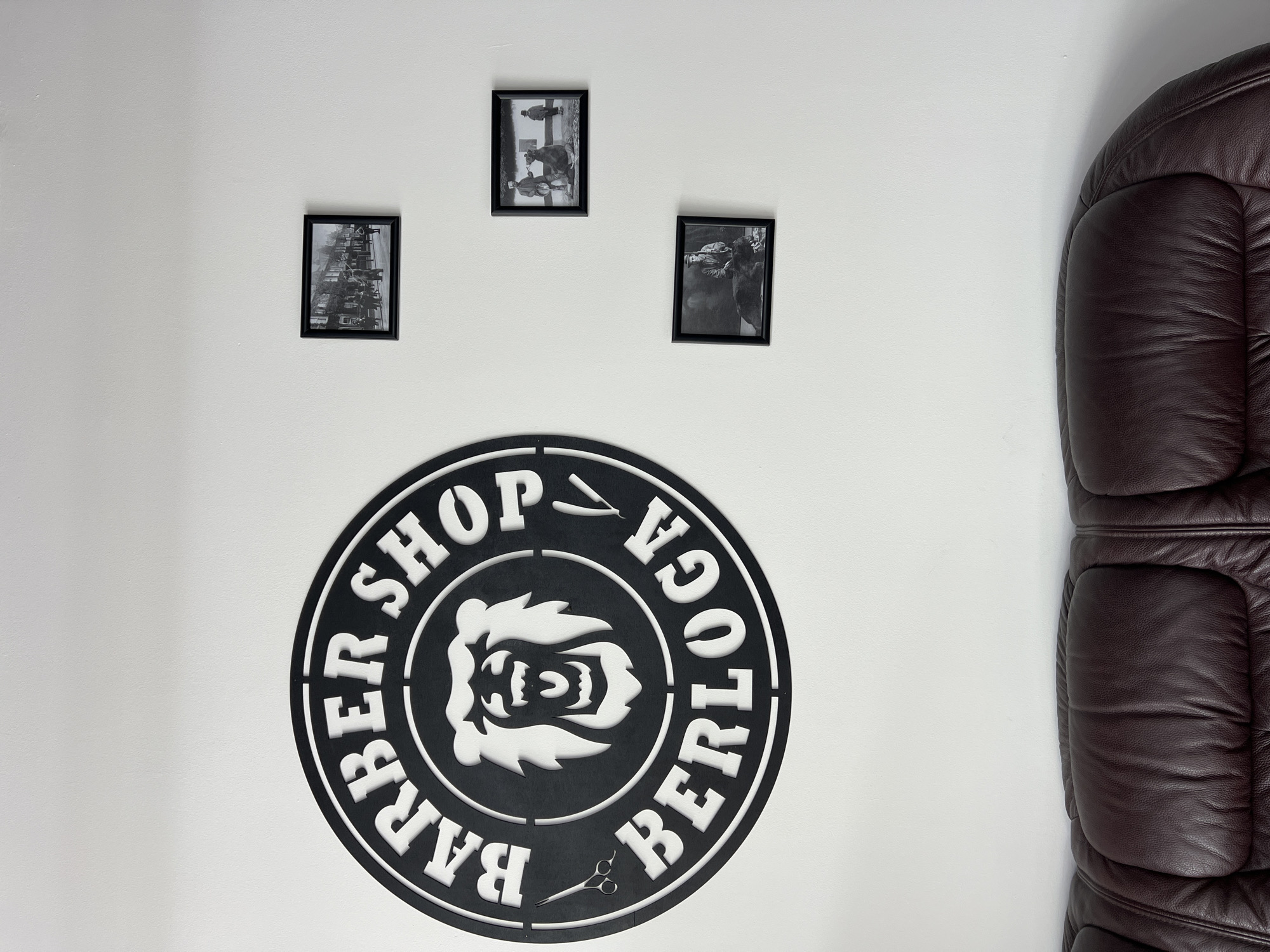 A wall is seen in an indoor setting. Three black-framed photographs are arranged near the top, depicting grayscale images. Below the photographs, a black circular sign with white lettering and a design element takes up a significant portion of the wall space. The sign reads "BARBER SHOP BERLOGA," and contains a stylized animal face, a straight razor, and a pair of scissors. To the right, a leather chair is visible, partially cut off by the frame.
There is no information in the image to suggest this is Ternopil, Ukraine.
