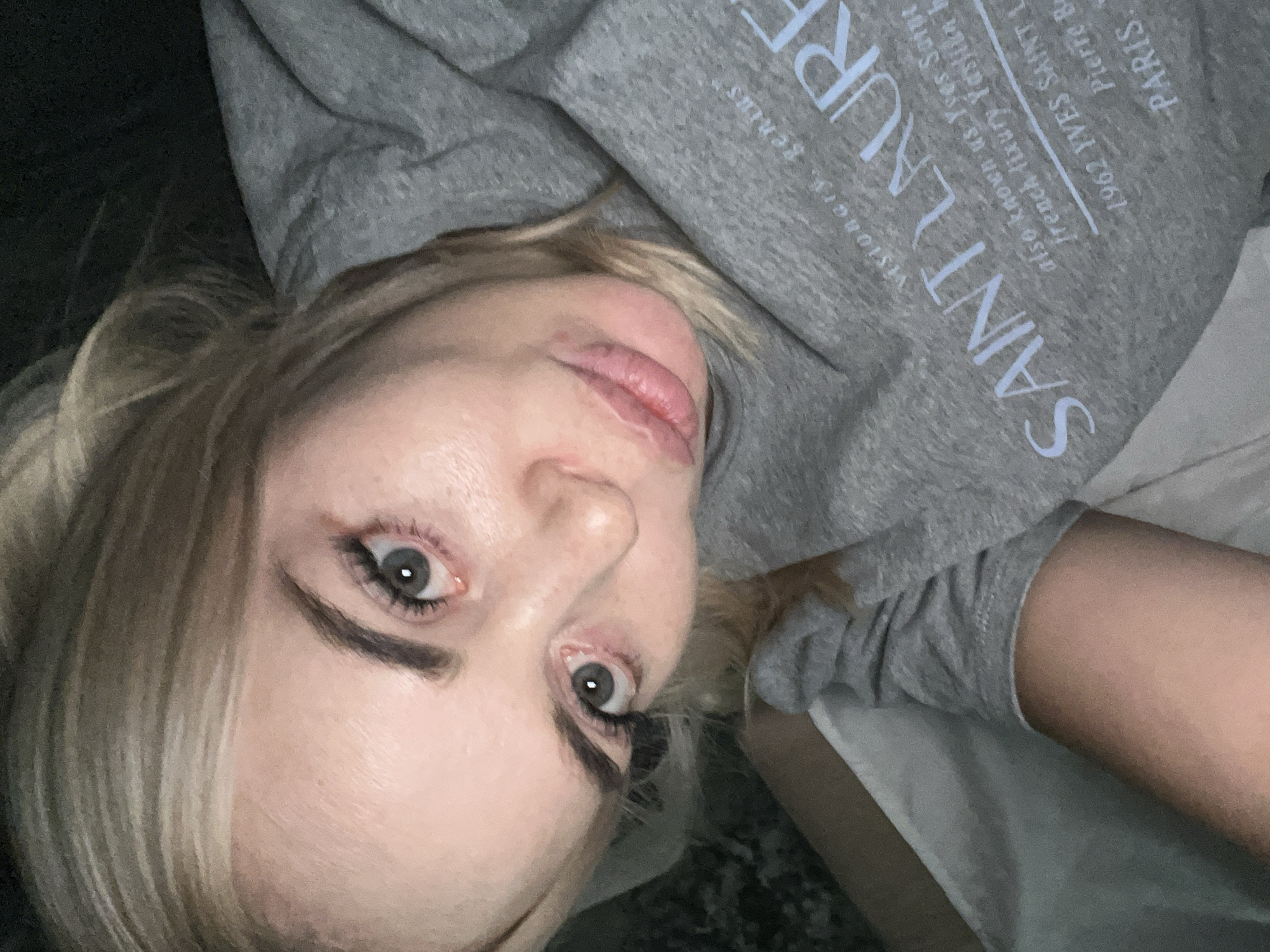 A close-up, overhead shot shows a blonde woman lying down in bed, looking at the camera. Her eyes are open, and she has makeup on. She is wearing a gray hoodie with "SAINT LAURENT" printed on it in white lettering. The woman is in a bedroom, with sheets visible in the background.