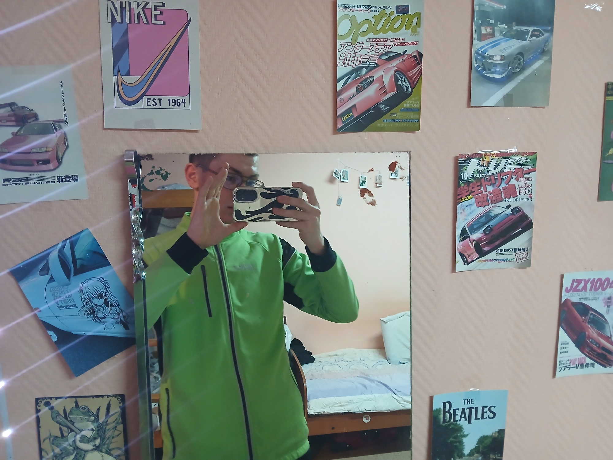 A man in a green jacket is taking a mirror selfie in a bedroom. The wall behind the mirror is decorated with various posters, including one of a pink Nissan R32 Skyline, a Nike poster, a Japanese car magazine, a poster of a silver Nissan Skyline GT-R, another car magazine, a poster of a car with an anime girl on it, a frog poster, and a "The Beatles" poster. The man is holding a phone with a black and white case. A bed is visible in the mirror's reflection. The room appears to be in a residential setting. The location is Chaikovsky, Russia.