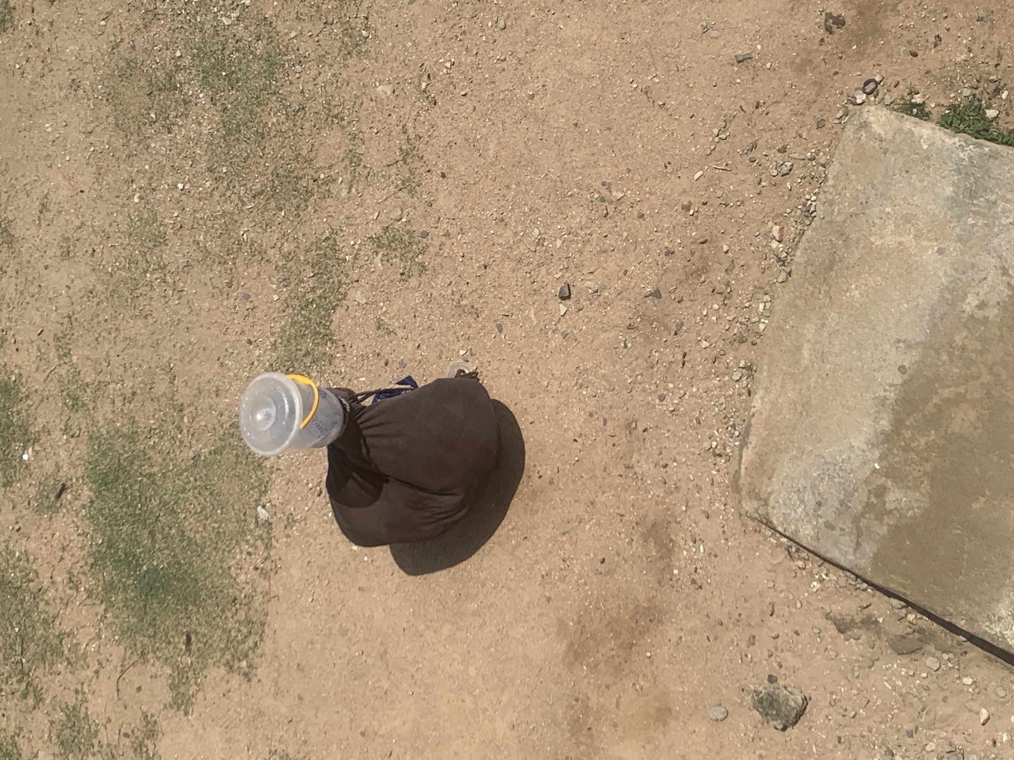 A person wearing dark clothing is seated on the ground, with a clear plastic bucket with a yellow lid on their head. The person's legs are tucked beneath them, and they are facing away from the camera. The ground is dry and sandy, with patches of sparse green vegetation and scattered pebbles. A large, flat concrete slab is visible in the lower right corner of the image. The scene appears to be in an outdoor, possibly rural or semi-urban, environment. The location is identified as Jalingo, Nigeria.