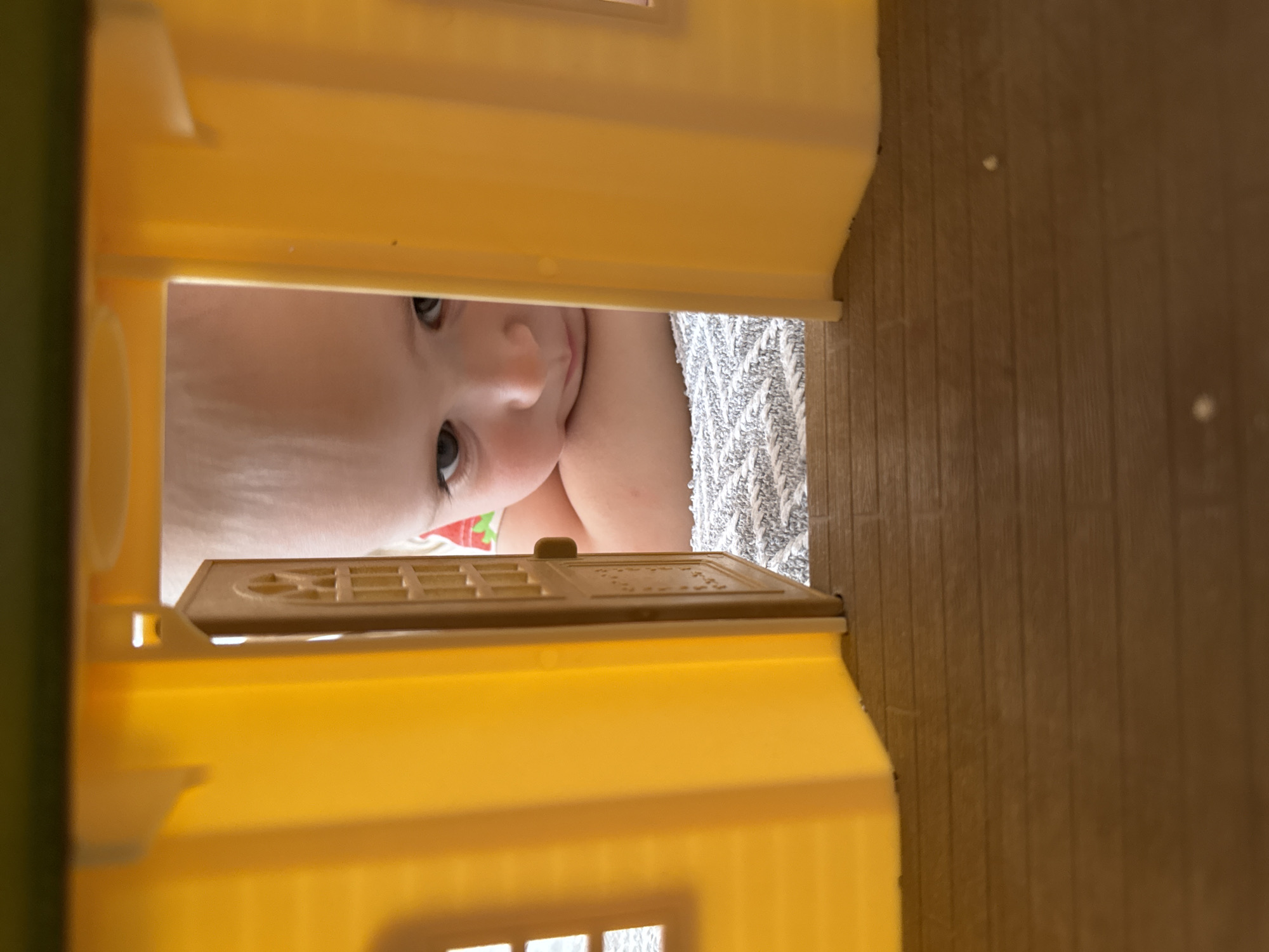 A close-up, vertical shot shows a baby looking into a yellow dollhouse. The baby's face, with light skin and dark eyes, is positioned in the center, peering through the doorway of the dollhouse. The dollhouse is made of yellow plastic, with visible windows and a door frame. Behind the baby, a gray and white textured surface is visible, possibly a blanket or towel. To the right, a wood-grain surface suggests a floor. The image does not contain information indicating that the photo was taken in Chaikovsky, Russia.