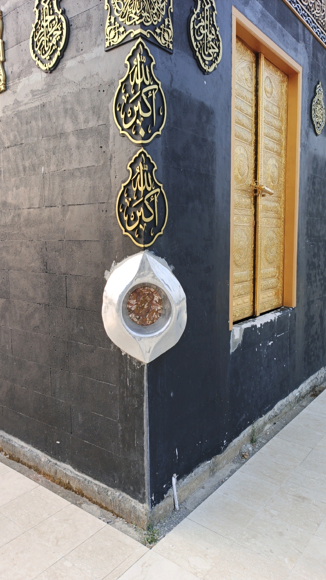 The image shows a close-up, exterior view of a corner of a dark grey, brick-textured building, possibly a religious structure, adorned with ornate gold calligraphy. A distinctive feature is a silver, tear-drop shaped encasement embedded in the corner, containing a circular, reddish-brown stone fragment. To the right of the encasement, a large, golden double door with intricate engravings is visible, set within a light brown frame. The building stands on a light-colored tiled floor, with a narrow strip of rough ground and some sparse vegetation along its base. The scene appears to be an outdoor location, potentially in the city of Campurdarat, Indonesia, given the provided context. There are no people visible in the image.