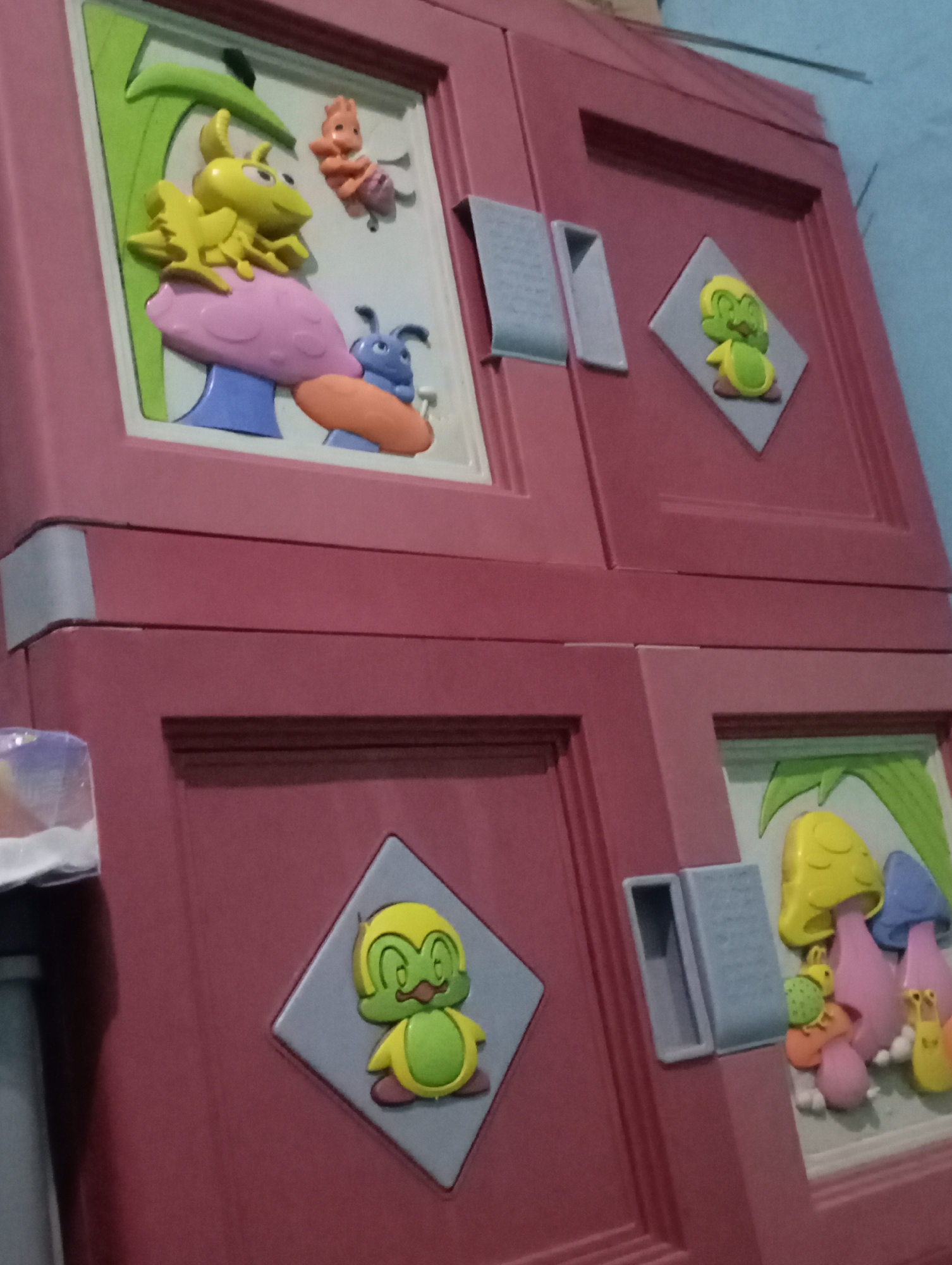This is a close-up, eye-level, medium shot of a red plastic toy cabinet with embossed cartoon characters. The cabinet is designed with a playful, nature-themed aesthetic, featuring various insects and mushrooms. The top left panel displays a green stalk, a yellow frog-like creature sitting on a pink mushroom, and two other small insect figures, one orange and one blue. The top right panel has a smaller, diamond-shaped inset with a green, frog-like character. The bottom right panel also features a diamond-shaped inset with a similar green, frog-like character, and a larger panel with several colorful mushrooms and small insect figures. The cabinet has grey handles that appear to be inset into the doors. The background is a plain, light blue wall. The image quality is slightly blurry, suggesting it may have been taken with a mobile phone camera. The scene is indoors and the lighting is moderate. The location is identified as Kecamatan Haurgeulis, Indonesia.