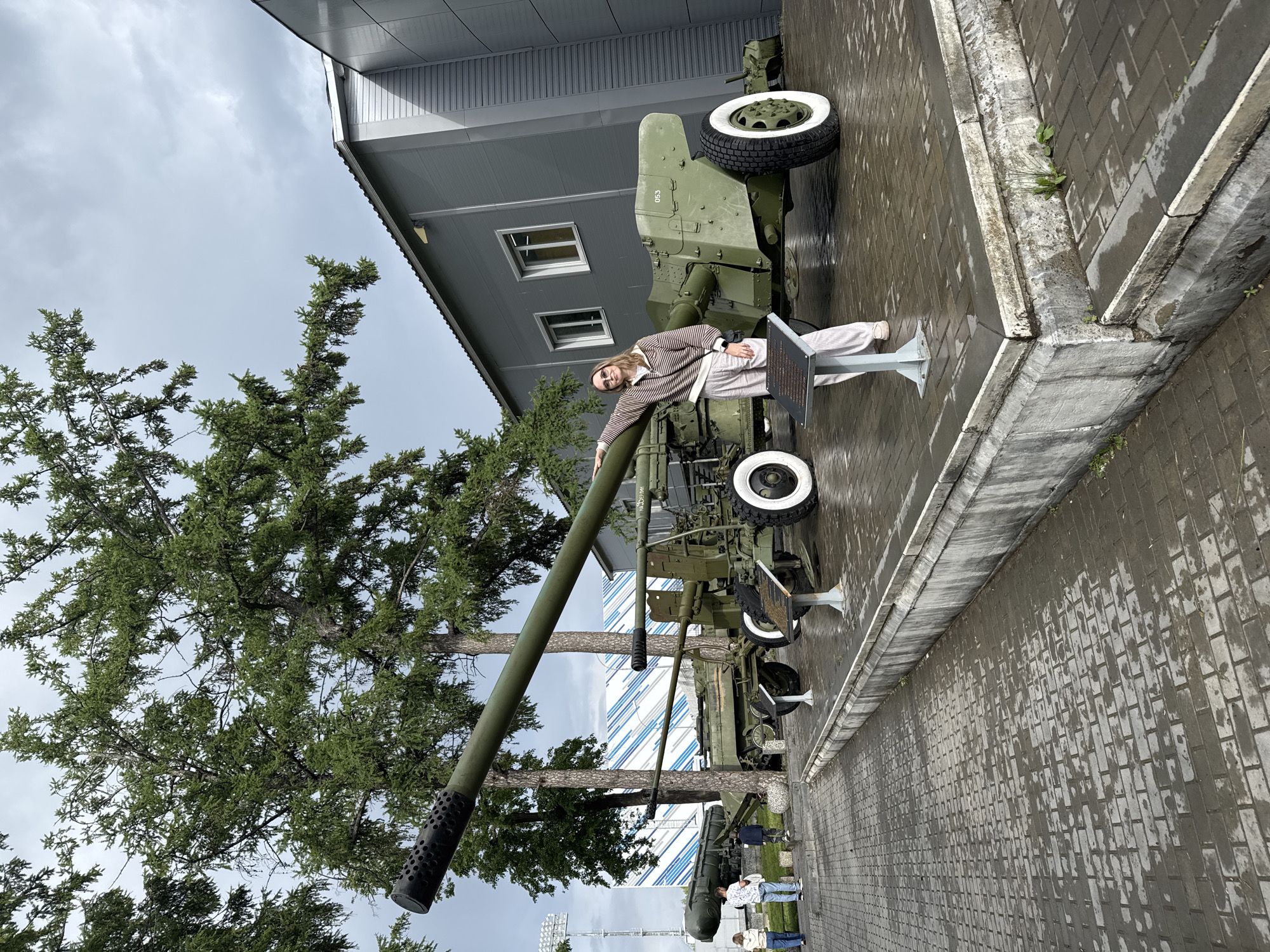 A woman is posing next to an anti-aircraft gun on display outdoors in Votkinsk, Russia. The scene features a large green anti-aircraft gun with white-rimmed wheels and a long barrel pointing upwards. A woman with sunglasses, wearing a striped shirt and light pants, leans against the barrel, seemingly posing for a photograph. The gun is mounted on a low display platform. Behind the gun and the woman stands a building with a light gray facade and two windows. A tall green tree is positioned to the left of the gun, partially obscuring the sky, which is overcast with gray clouds. The ground is covered with wet paving stones. A few people can be seen in the distance behind the gun.