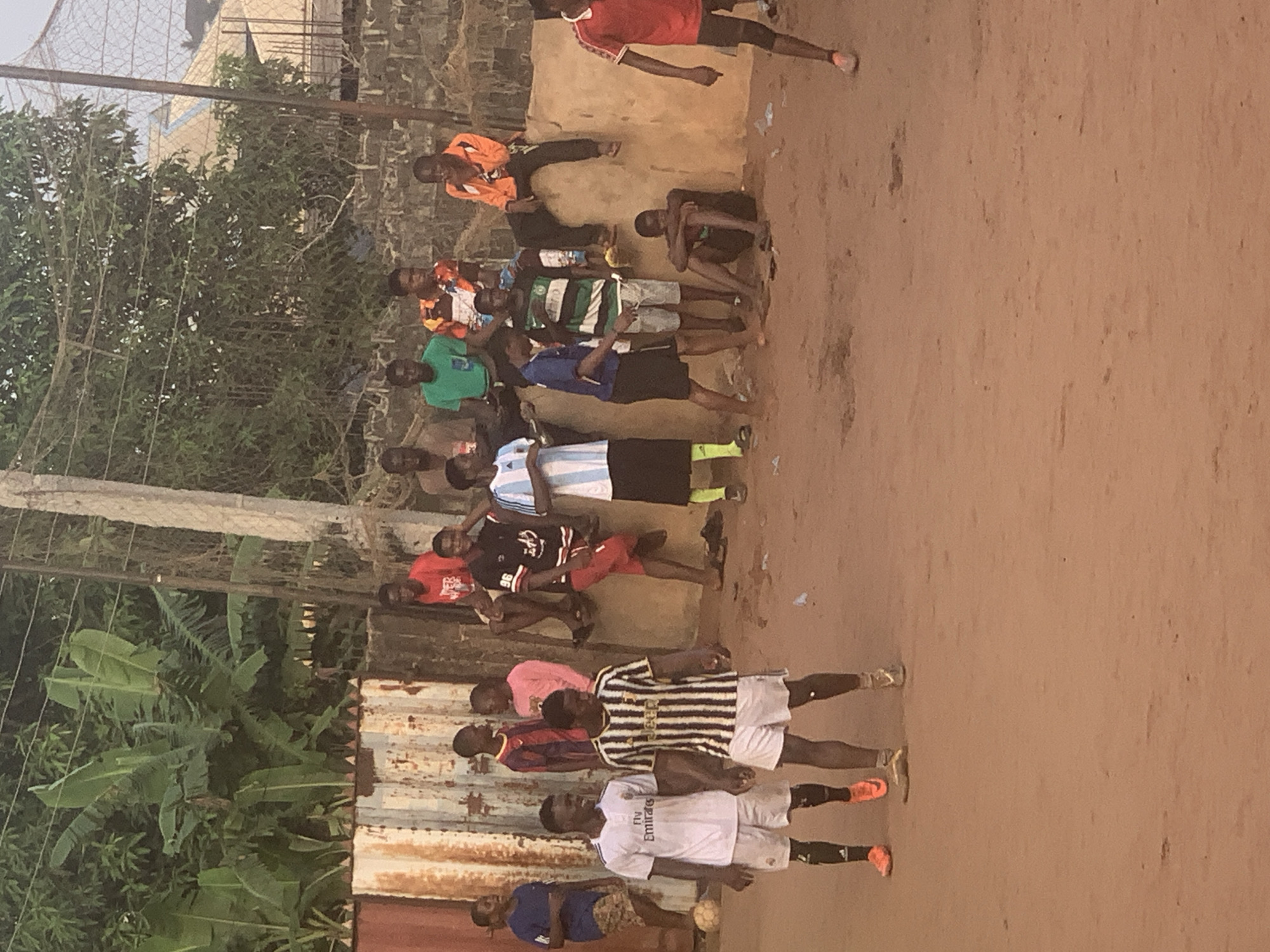 A group of approximately 15-20 young males, likely teenagers and young adults, are gathered outdoors on an unpaved, reddish-brown dirt ground in Onitsha, Nigeria. The image appears to be taken in the late afternoon or early evening, characterized by soft, warm light.
The subjects are dressed in a variety of casual attire and replica football (soccer) jerseys. Notable jerseys include a white one with "Fly Emirates," a black and white striped one bearing "JEEP," and a blue and white striped jersey resembling the Argentina national team's kit. One person wears a dark red jersey with the number "96." Most individuals are standing, while a few are seated on a raised dirt bank or on the ground. A white and black soccer ball rests near the feet of one individual in the foreground.
The setting appears to be an informal or makeshift football pitch. To the left, a corrugated metal fence separates the playing area from dense green vegetation, including large-leaved plants that resemble banana trees. In the upper left corner, a mesh net, possibly part of a goal or another fence, is visible. The overall scene suggests a community gathering, likely related to a football match or practice session. The expressions on the faces are generally relaxed, indicative of a casual social environment.